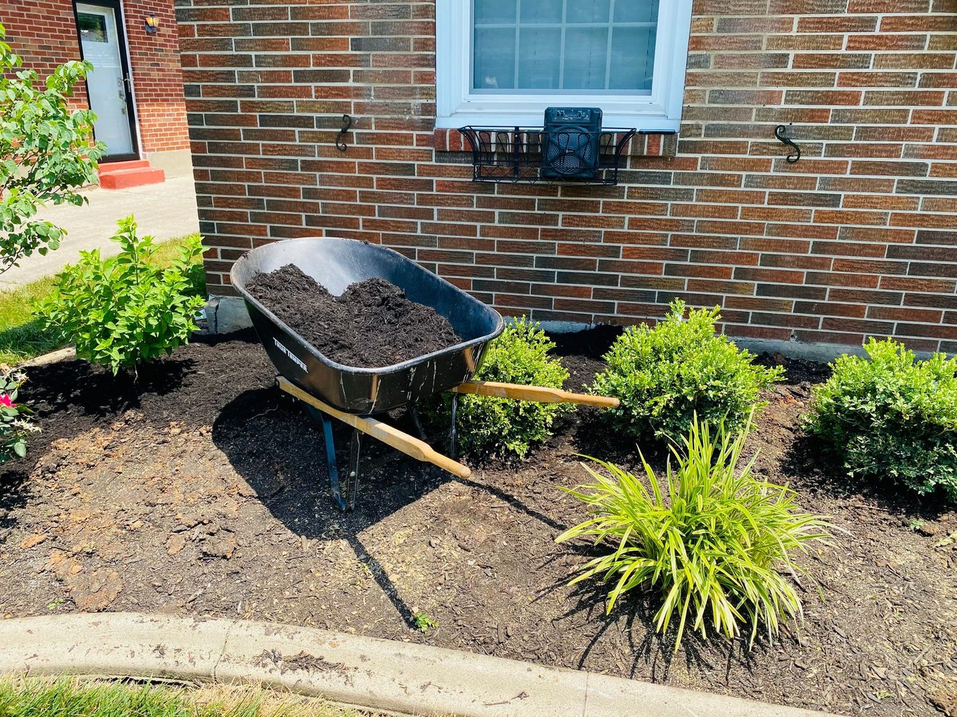 Wheelbarrow filled with dark mulch in a garden bed near a brick building.