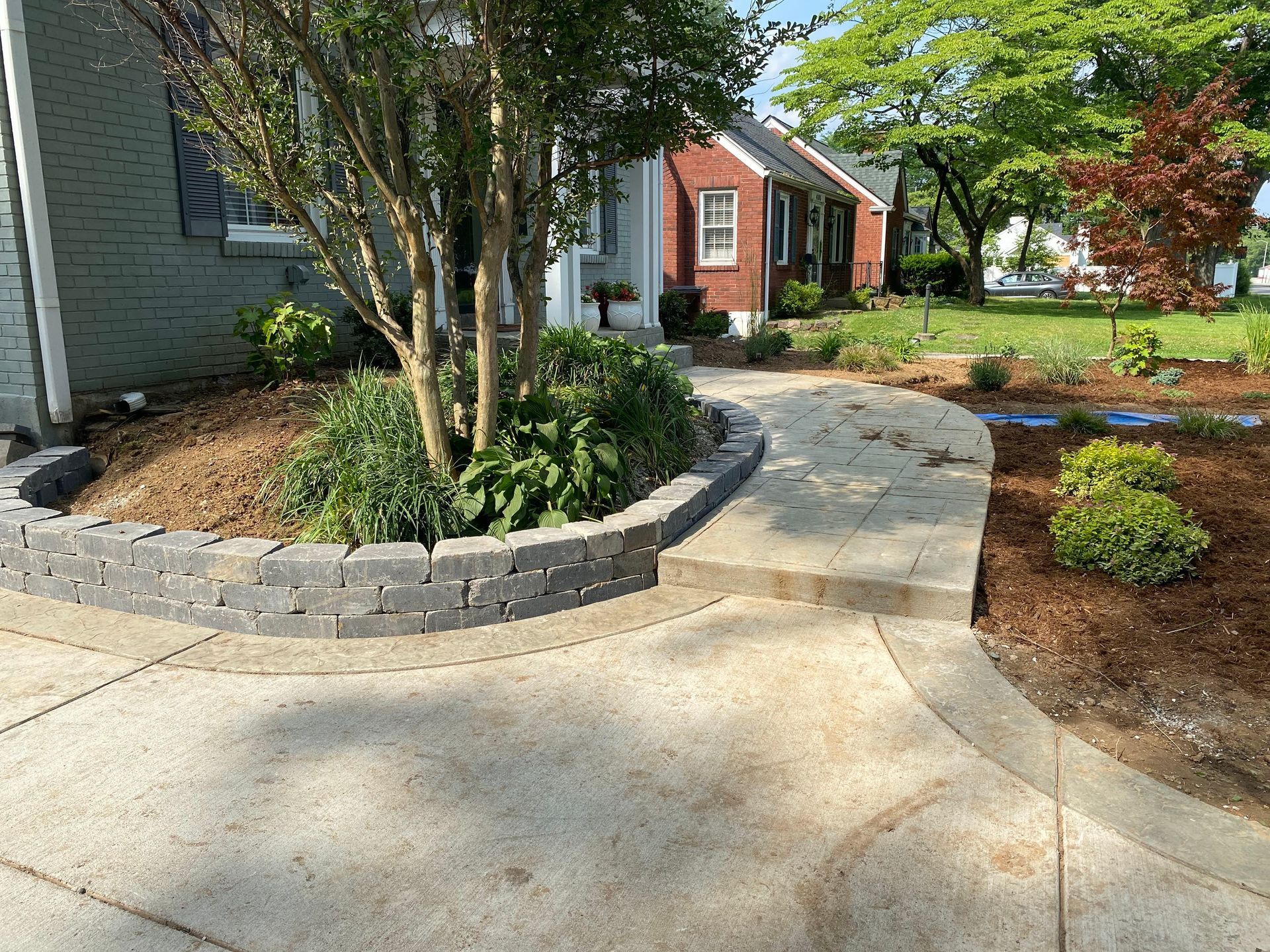 A concrete walkway curves past a landscaped bed with a retaining wall, leading to a brick house.