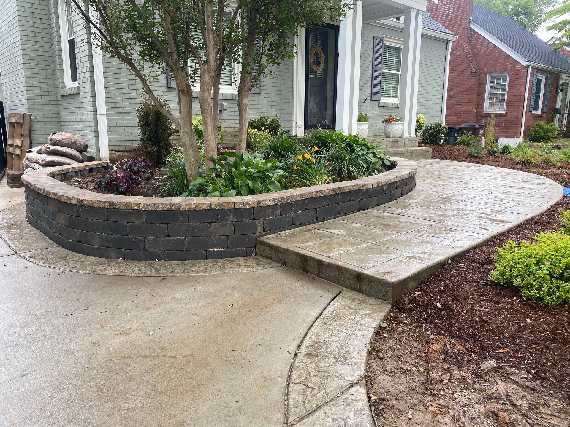 A concrete walkway and brick planter with a tree, flowers, and a house in the background.