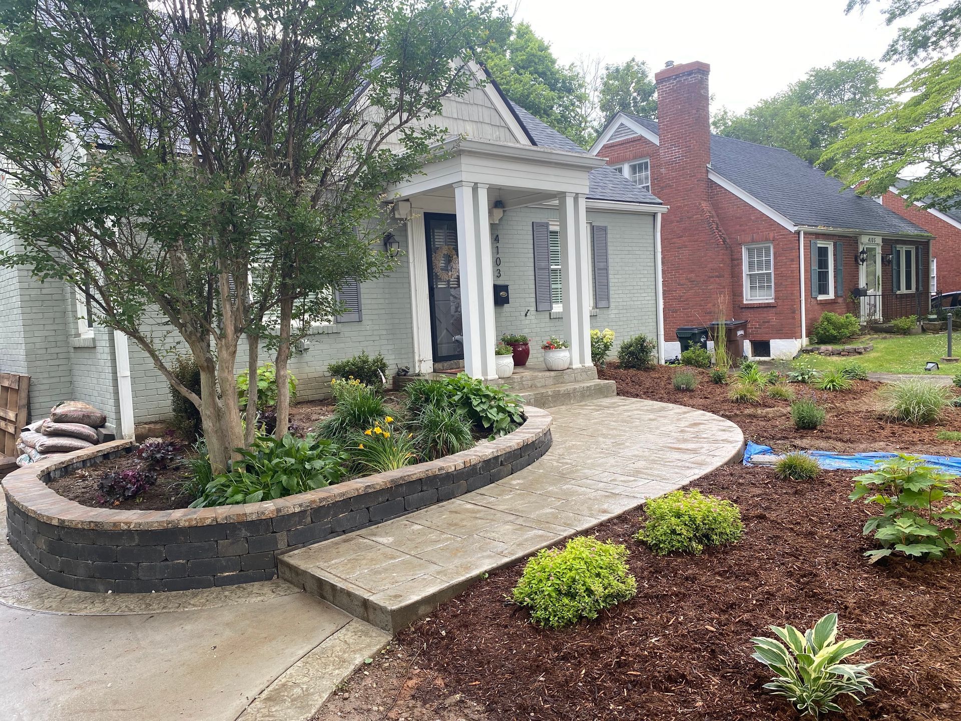 A light green house with a brick walkway and garden, trees, and flowers.