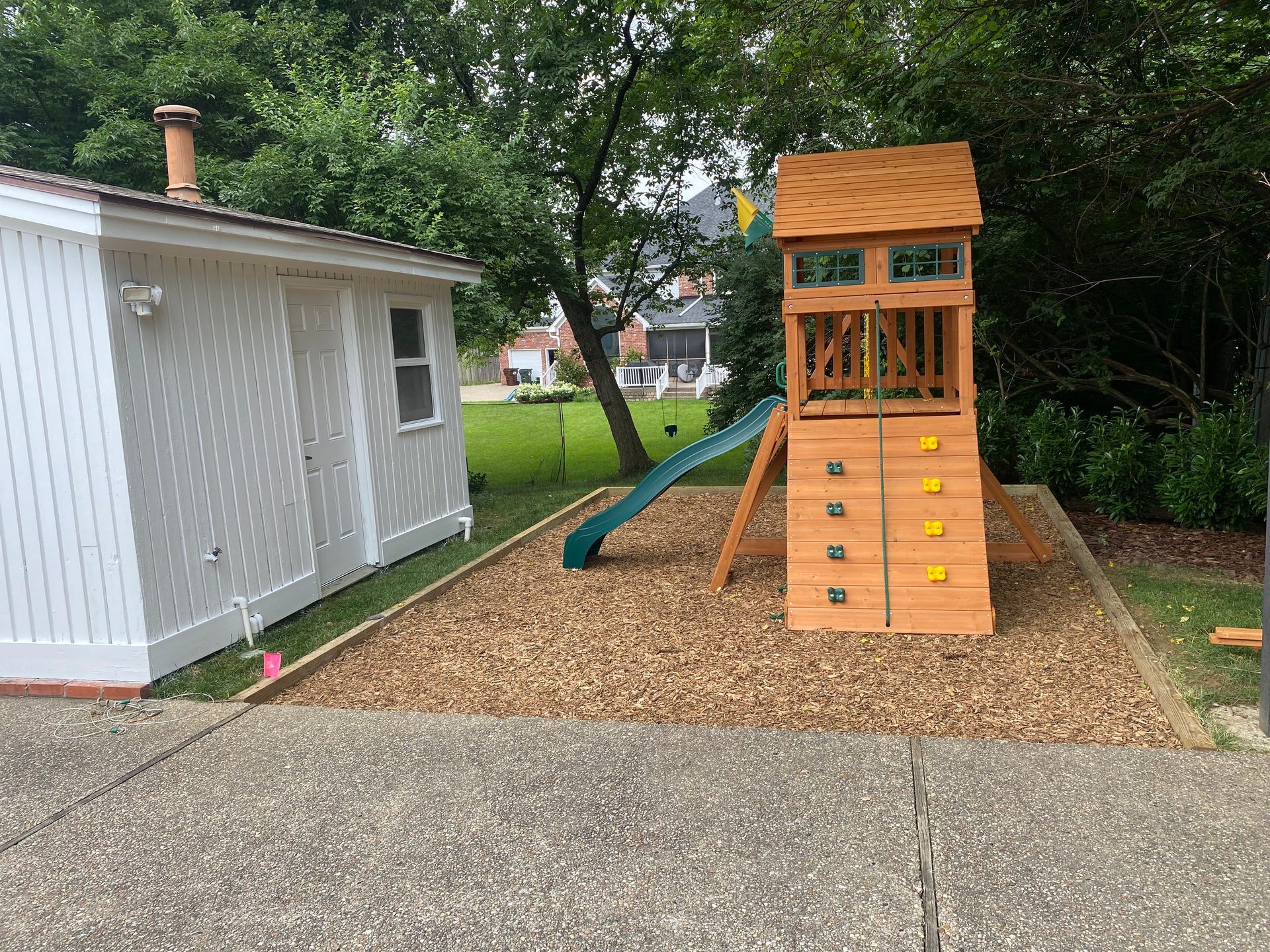 Play set with slide and climbing wall in a yard with wood chips, next to a white shed.