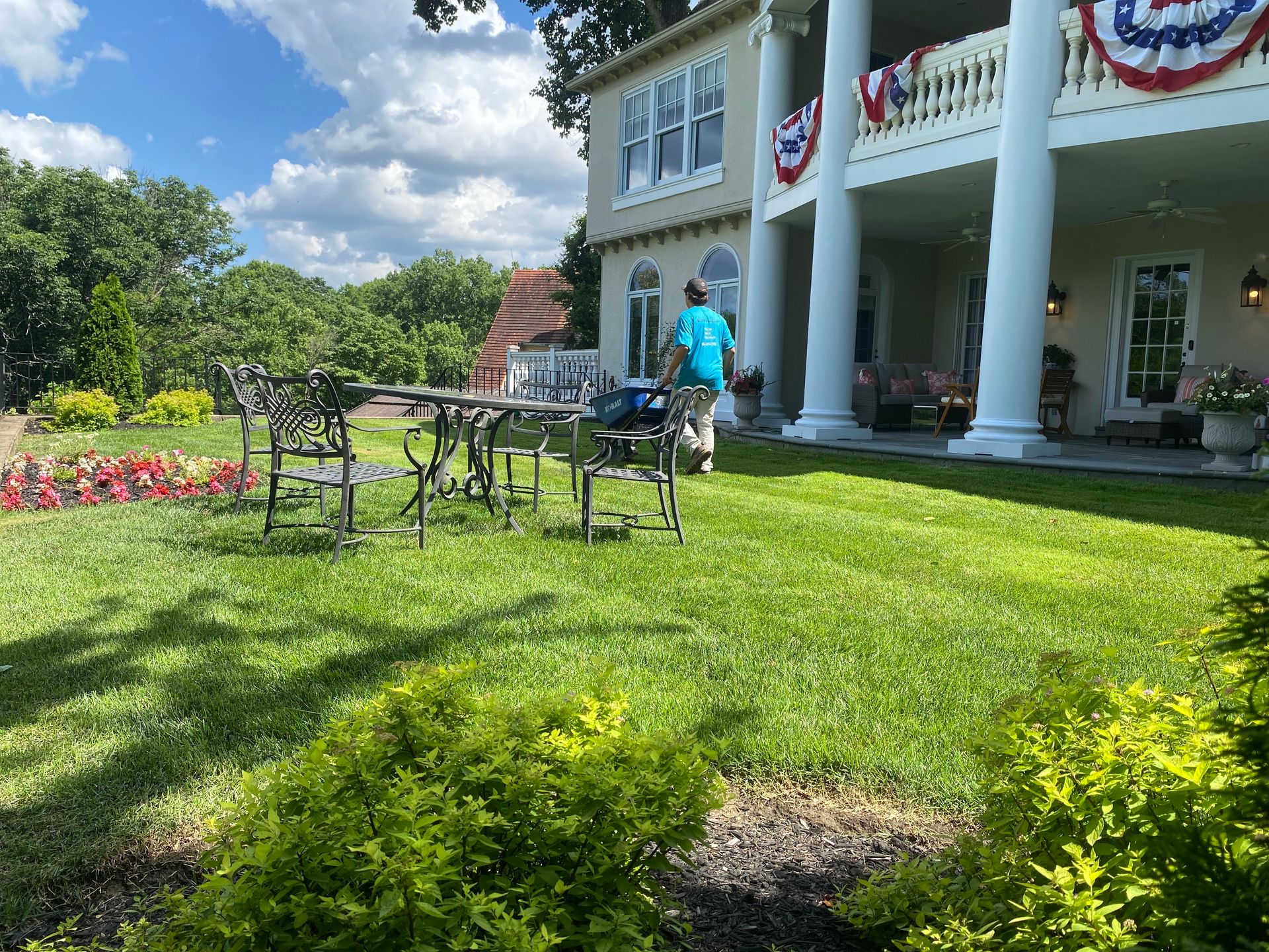 Man on lawn near a white house with a large porch, red, white, and blue decorations, and a wrought-iron table and chairs.