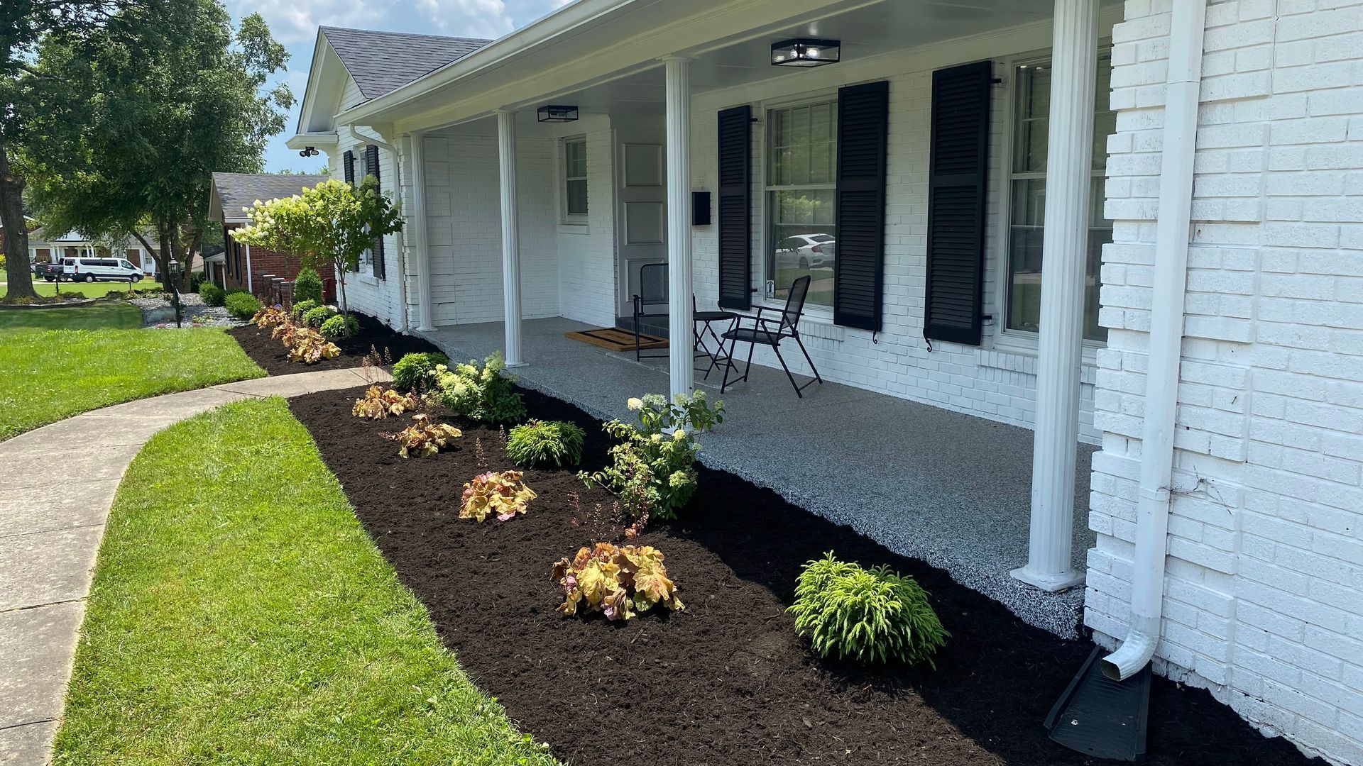 A white-painted brick house with black shutters and a porch, bordered by a mulch bed and walkway.