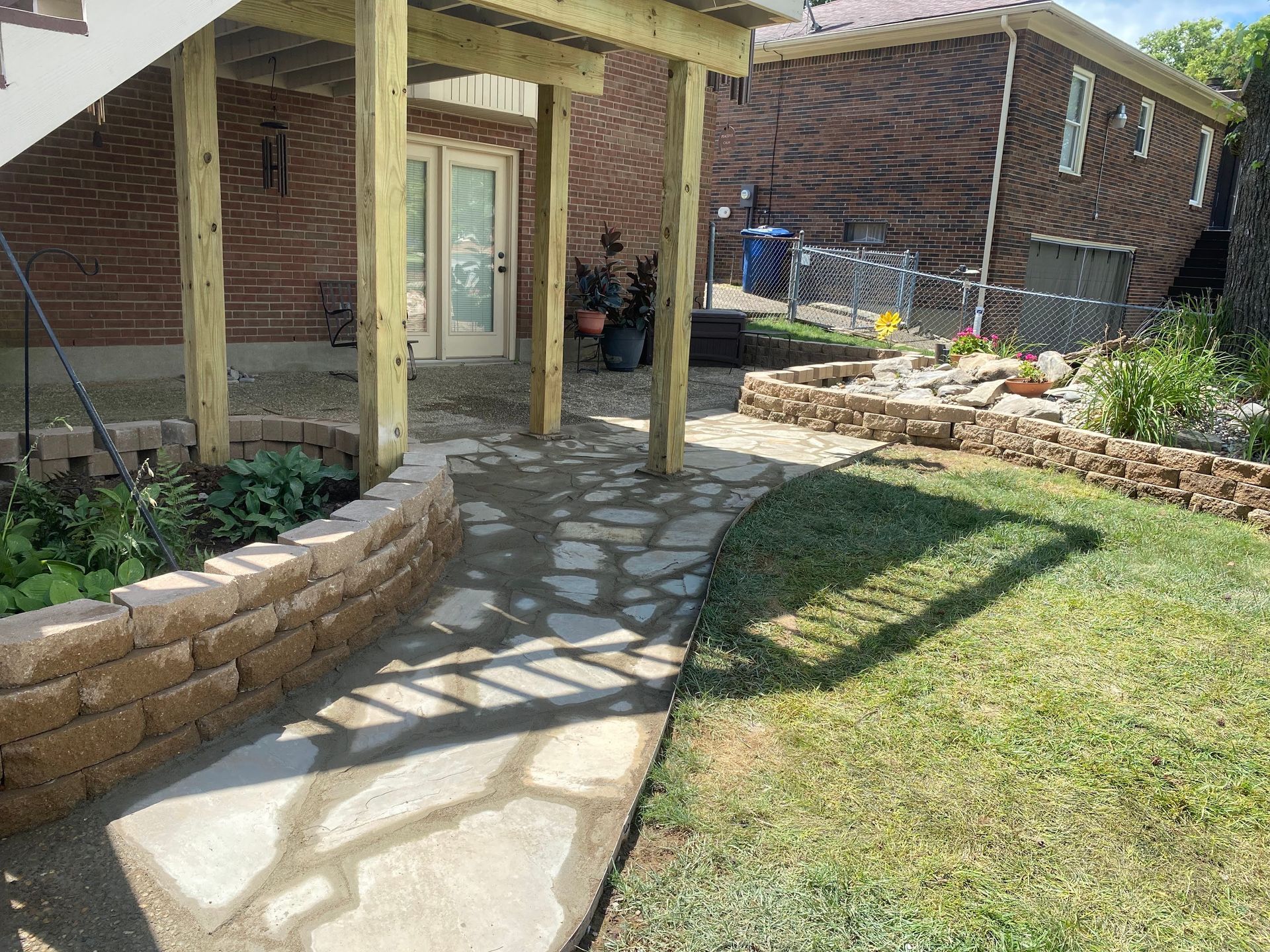 Stone pathway leads to a back door beneath a deck; a low brick wall borders a garden on the left, grass on the right.