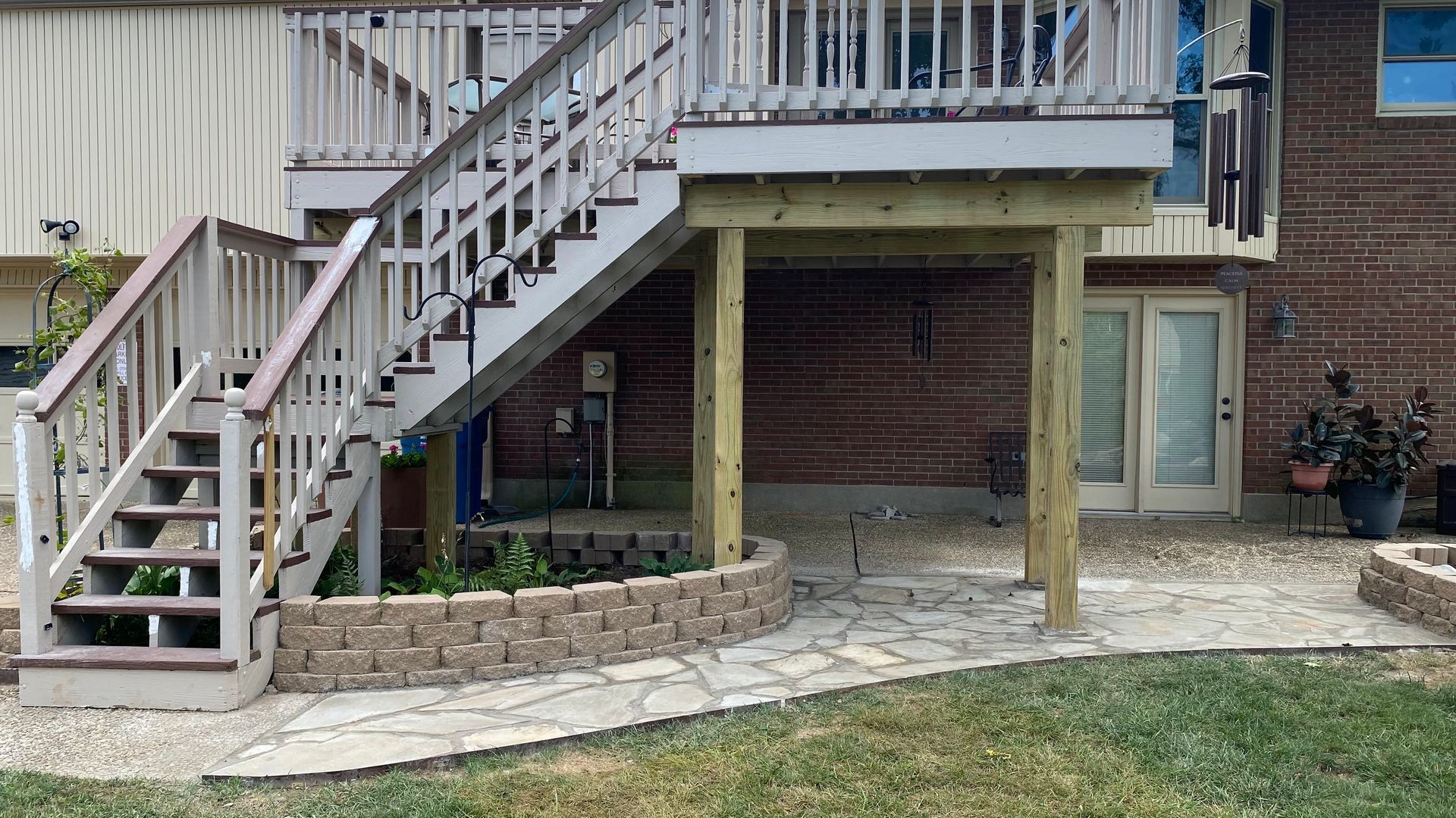 Wooden deck with stairs, brick house background, stone patio, and small garden bed.
