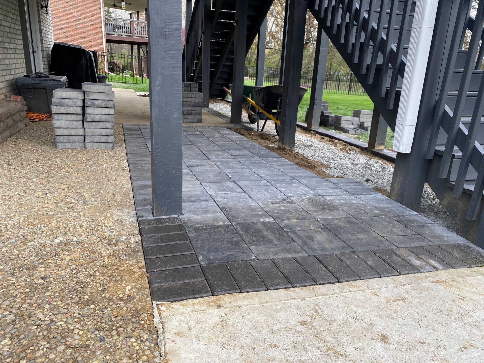 Patio area with dark grey pavers being installed under a black staircase, next to a gravel path.