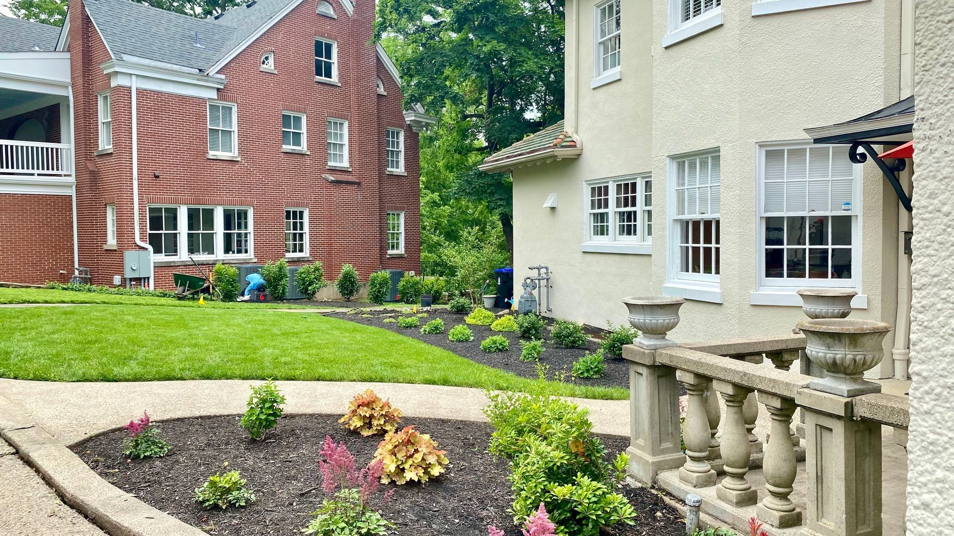 Two houses with lush landscaping. One is red brick, the other stucco. A stone patio is in the foreground.