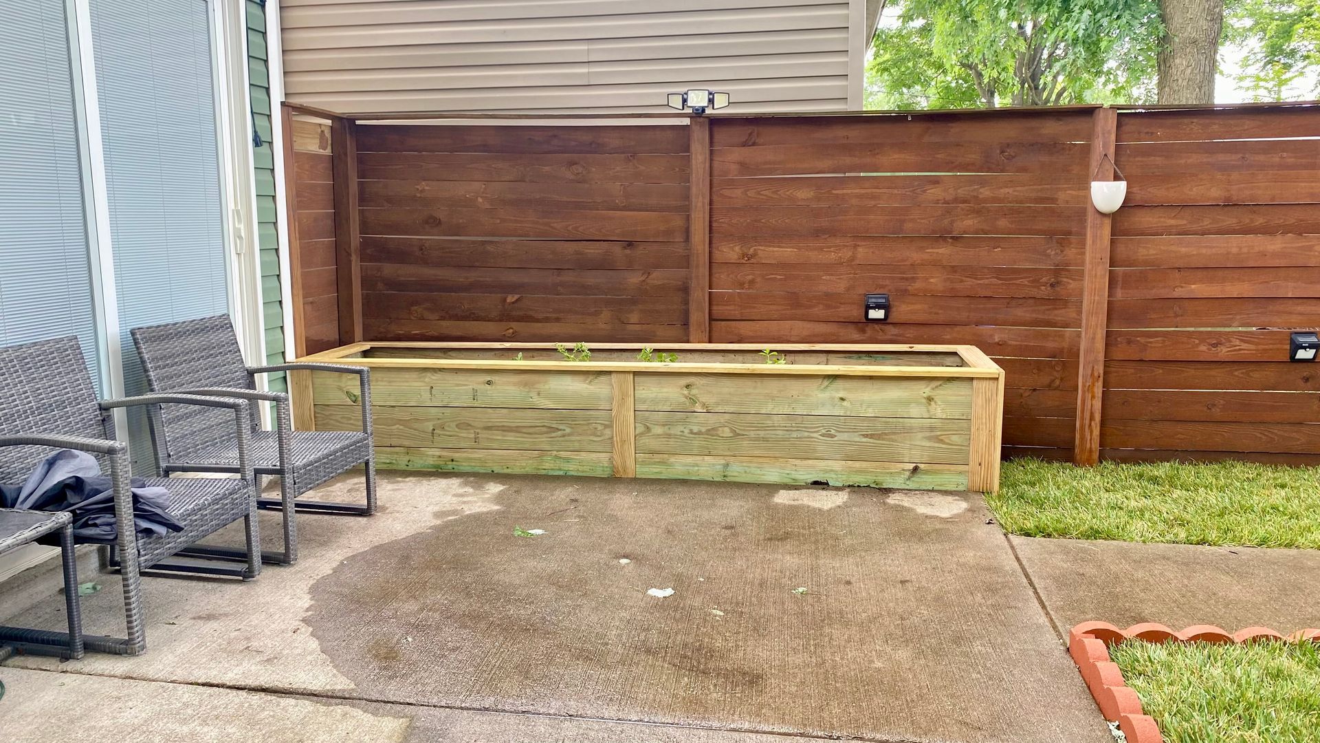 A wooden raised garden bed sits against a brown fence on a concrete patio, with two chairs to the left.