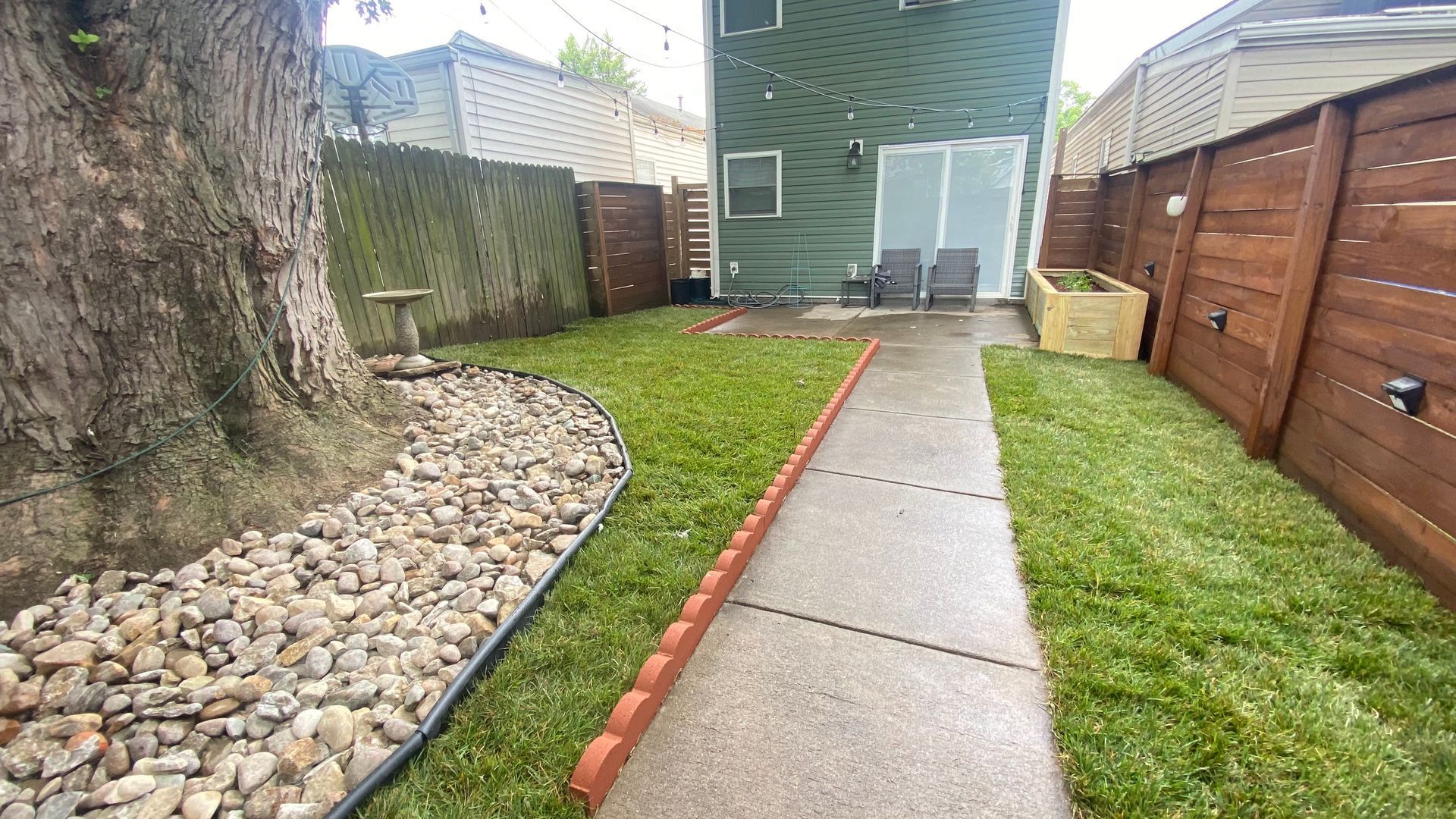 Backyard with a stone-lined tree, green grass, a concrete path, and wooden fences. A green house is in the background.