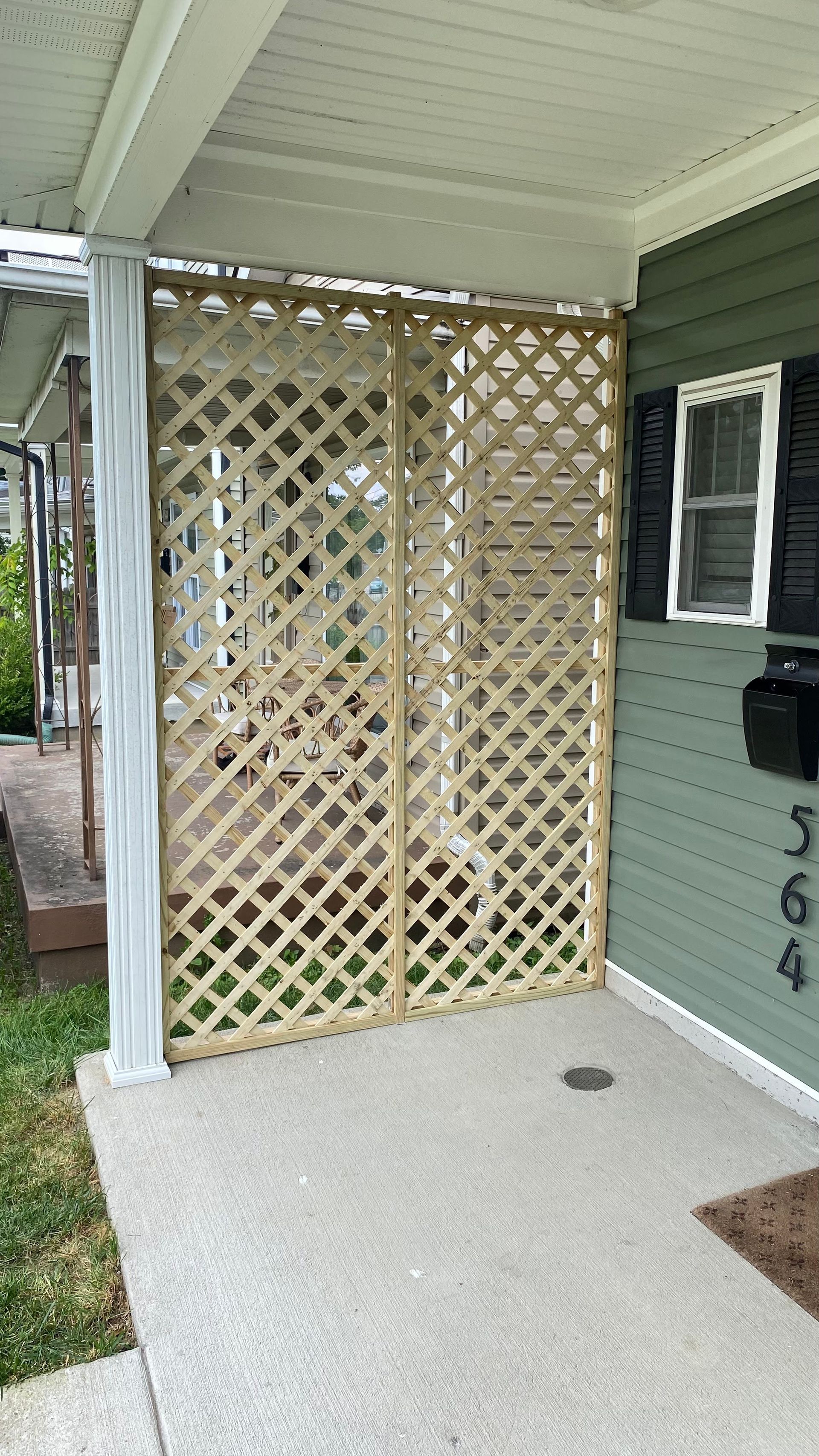Wooden lattice screen on a porch, separating the space. Green siding and concrete flooring, with a house number sign visible.