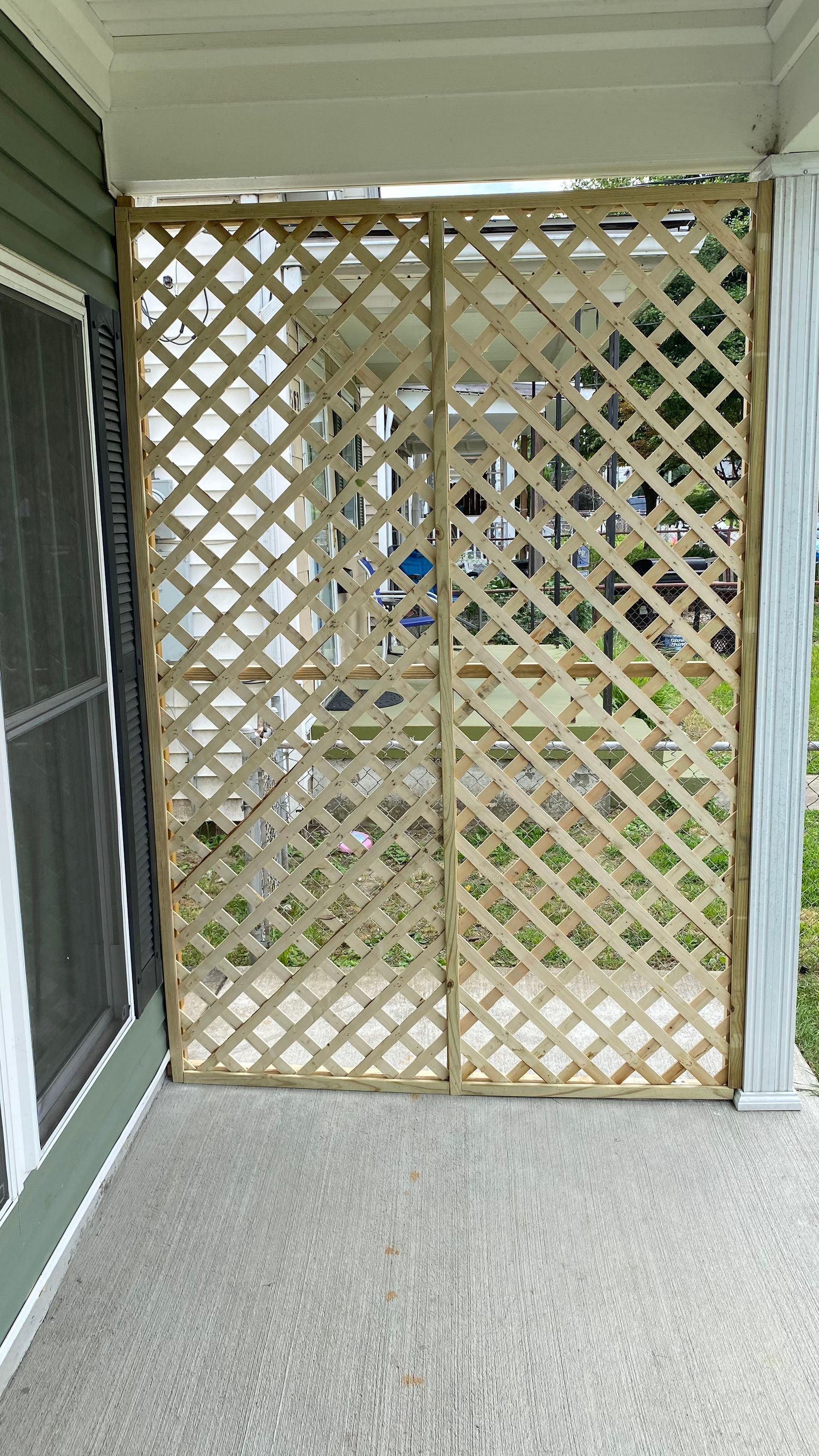 A light-colored wooden lattice screen on a porch, between a screened window and a white column.