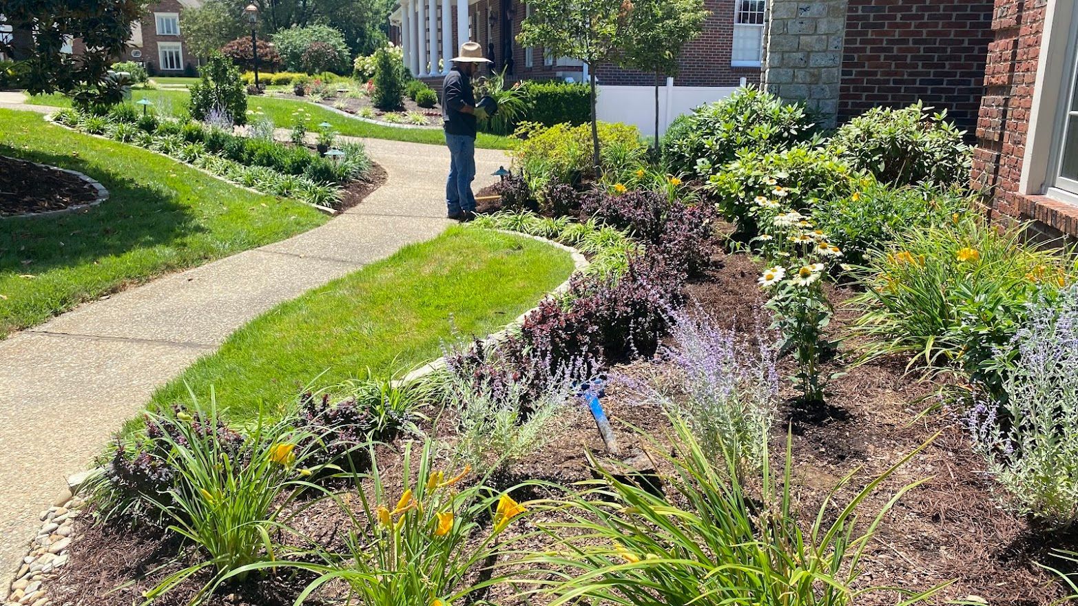 A person stands near a flowerbed with a stone path winding through a lush green lawn.