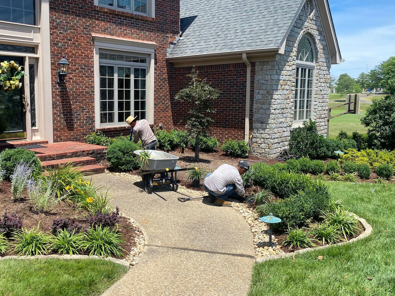 Two people tending to a landscaped yard in front of a brick and stone house; sunny day.