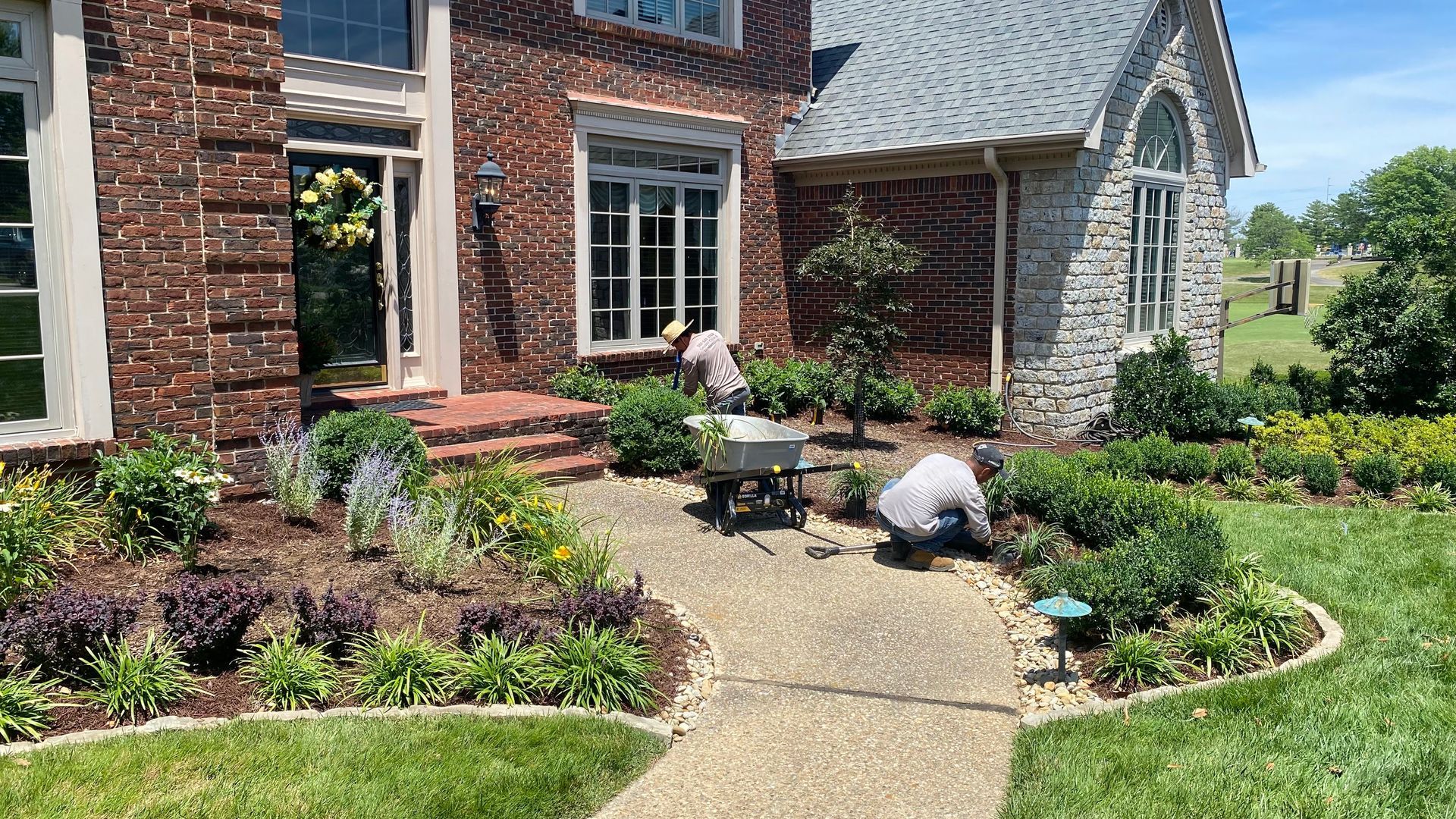 Two people landscaping flower beds in front of a brick and stone house. Green lawn and plants.