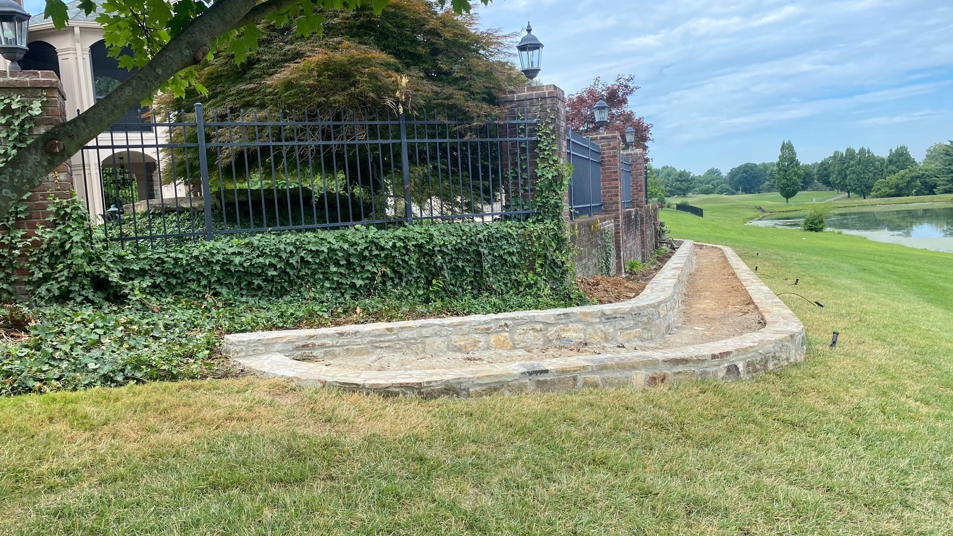Concrete edging borders a grassy area next to a lake, with a fence and building in the background.