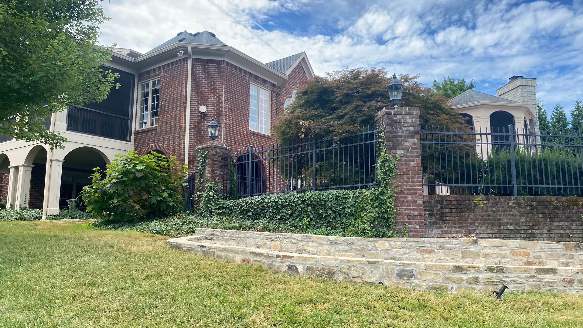 Large brick house with iron fence, stone steps, and green lawn under a blue sky.