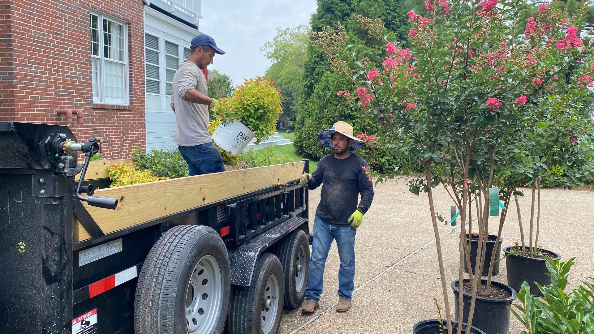 Two men loading a trailer with plants. One man is in the trailer; the other is outside the trailer near trees in pots.
