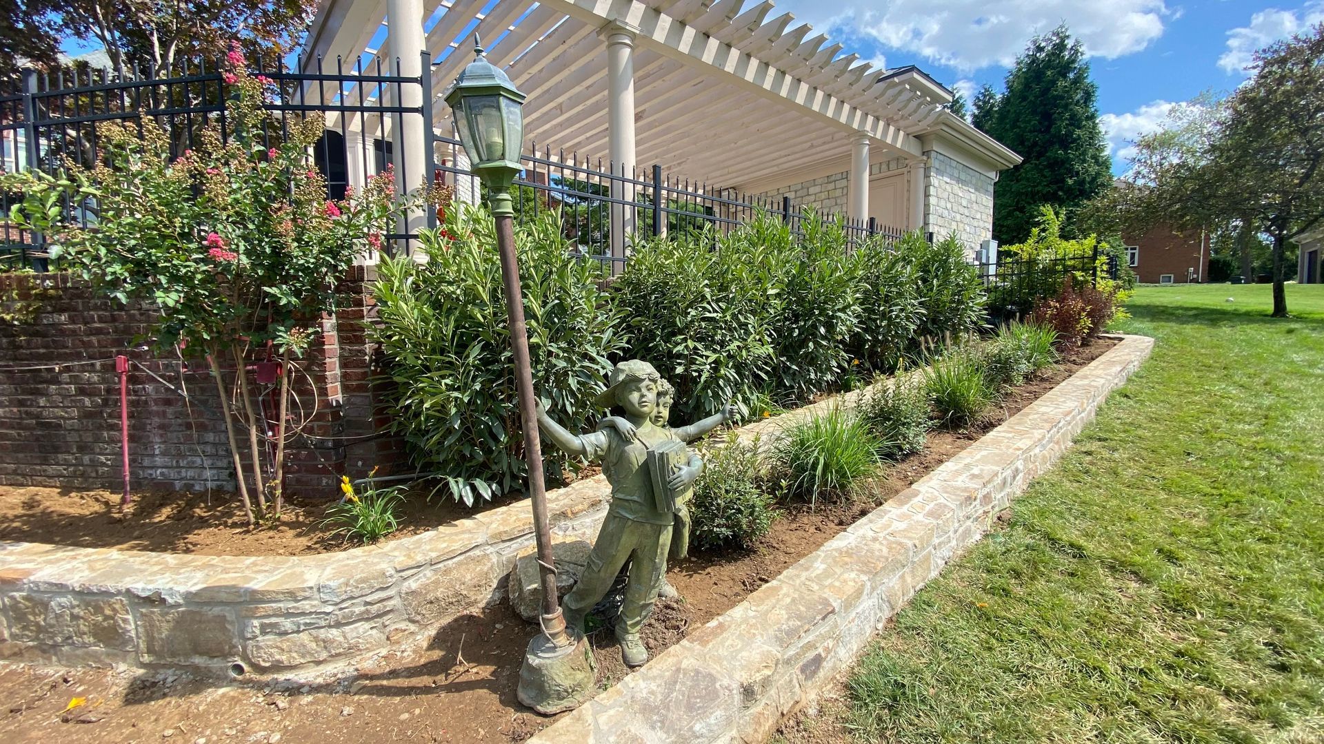 Statue of a child holding a street lamp in a garden bed near a building with a pergola.