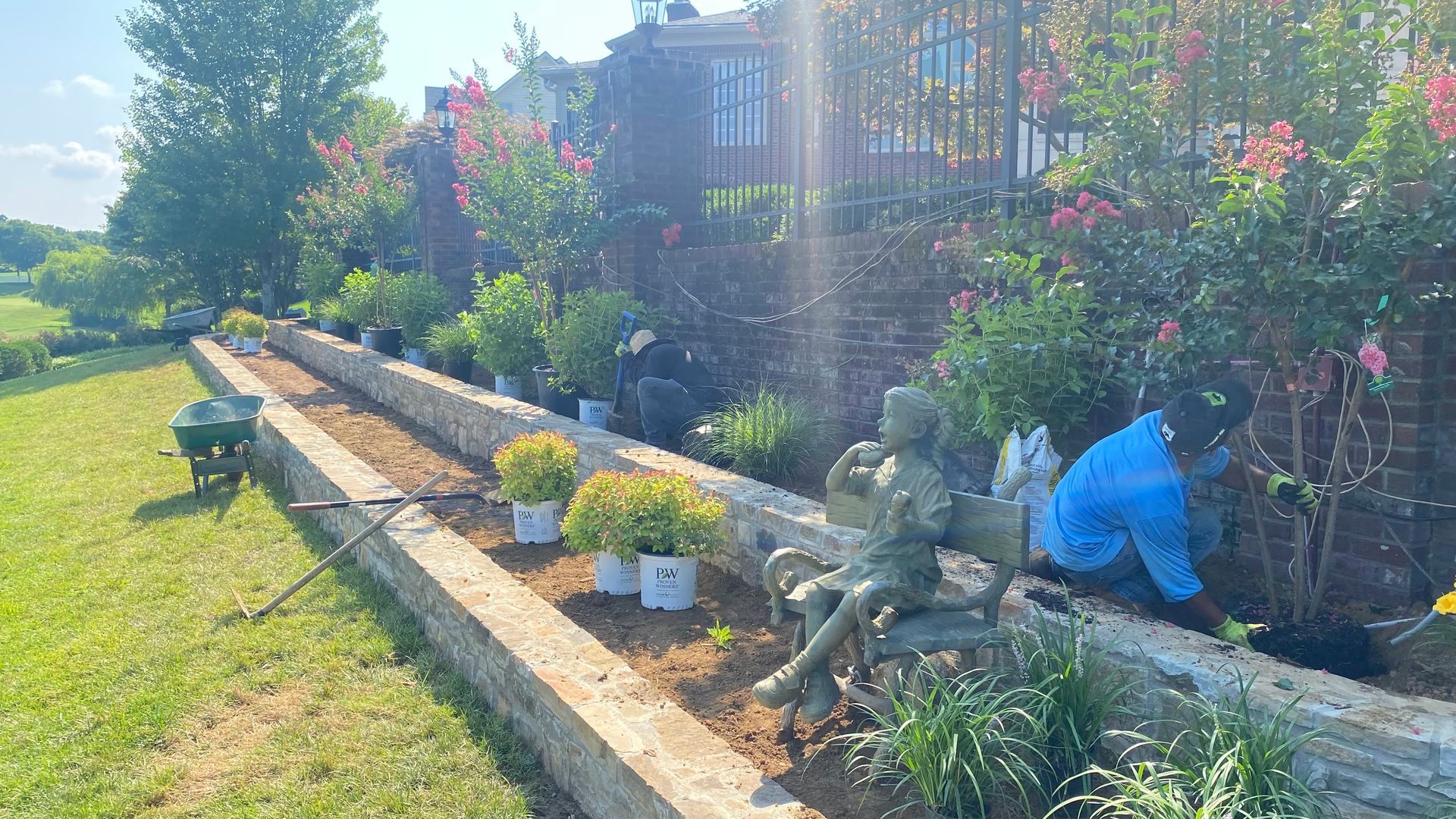 Person planting flowers in a garden bed along a brick wall, next to grass.