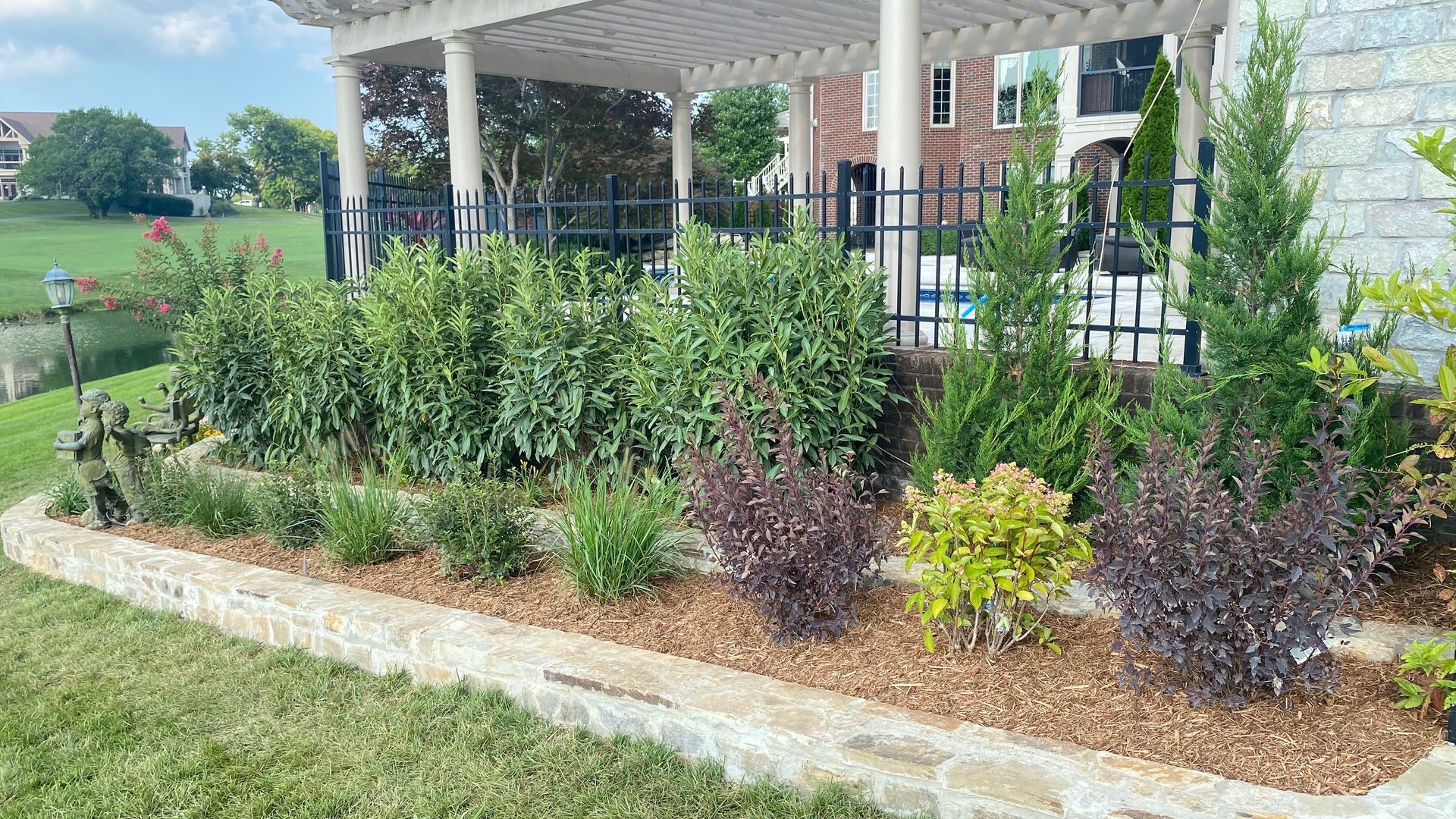 Stone-edged garden bed with varied shrubs, mulch, and pergola in background. Green grass and blue sky.