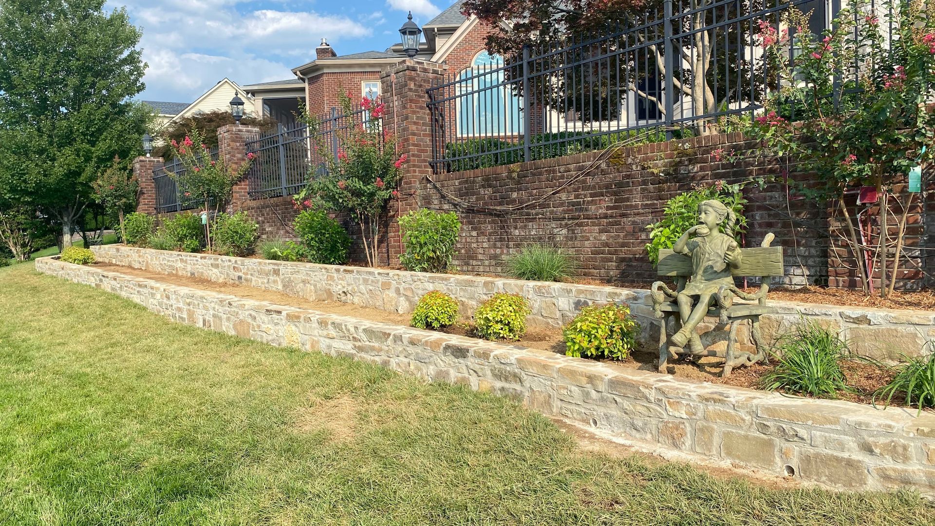 Brick building with stone tiered planters and a bronze statue on a green lawn.  The sky is blue.