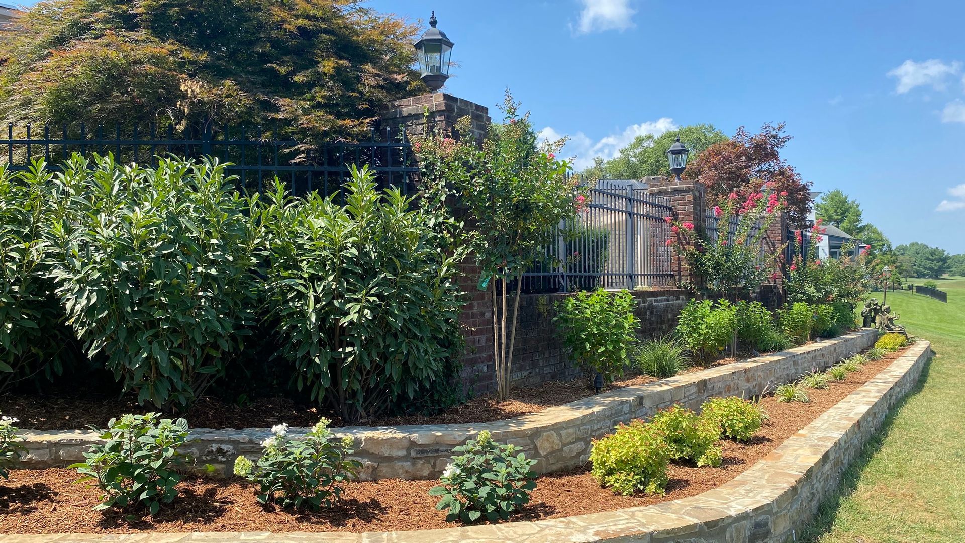 Stone retaining wall with a variety of shrubs and mulch beds against a backdrop of green foliage and blue sky.