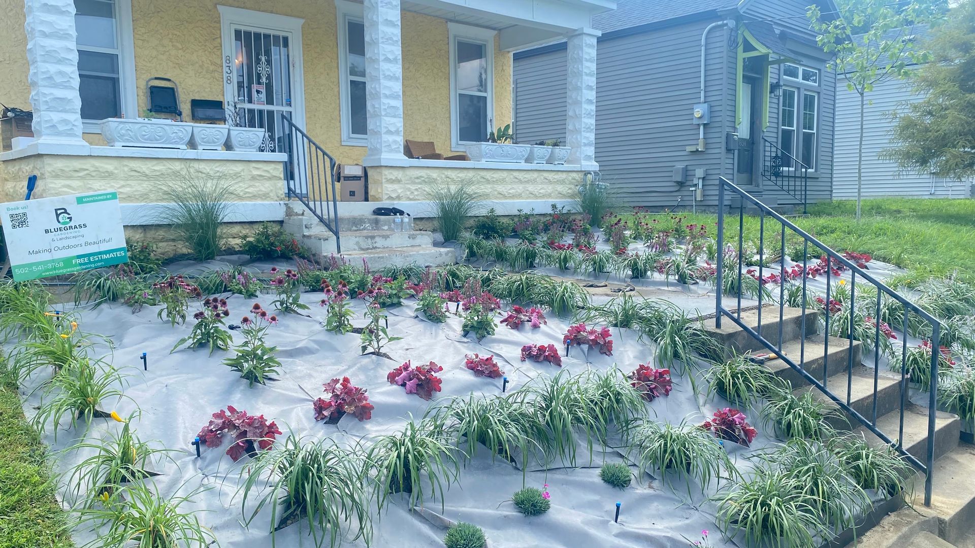 Yellow house with flowerbed, red flowers and green plants in front. Steps and railing lead up to the porch.