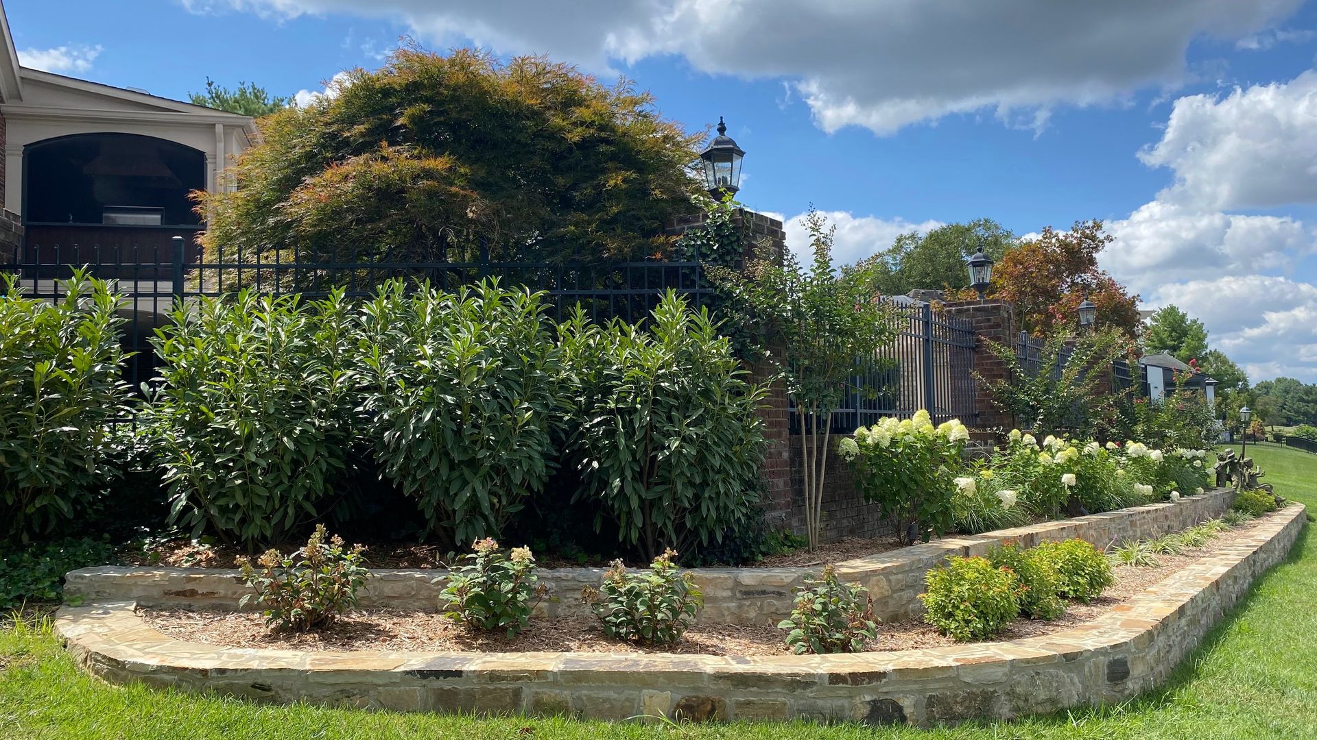 Raised garden bed with flowers in front of a house, under a partly cloudy sky.