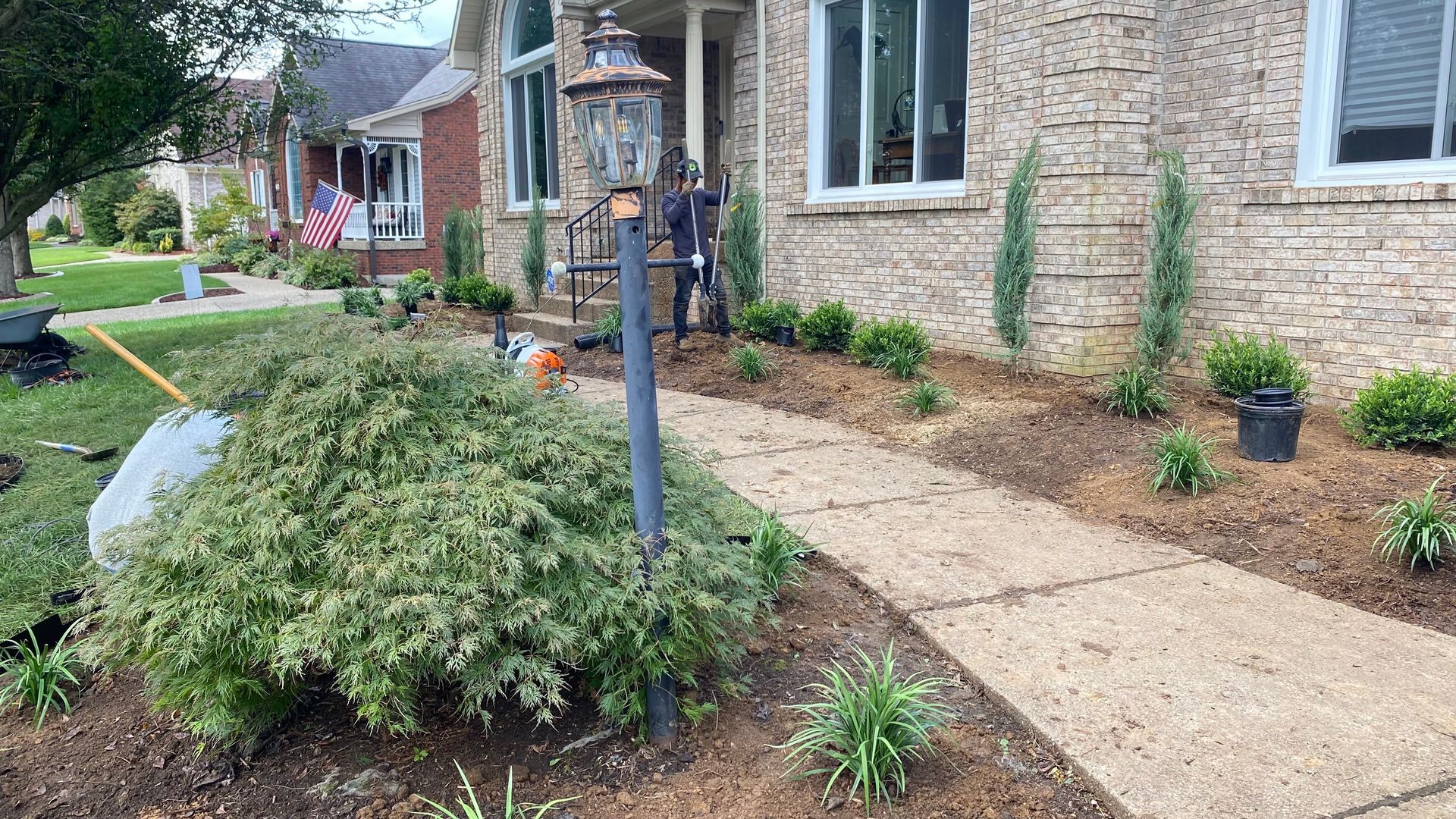 A person is landscaping a house's front yard with shrubs, a walkway, and a lamppost.