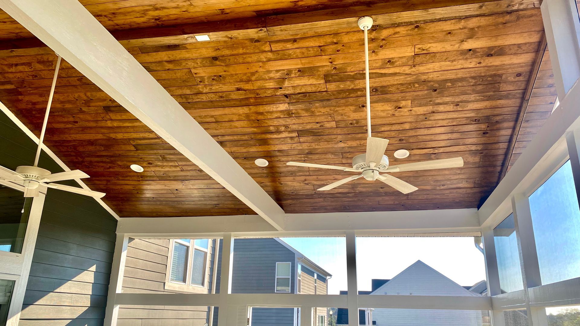 Screened porch with wood ceiling, white beams and two ceiling fans overlooking a suburban neighborhood.