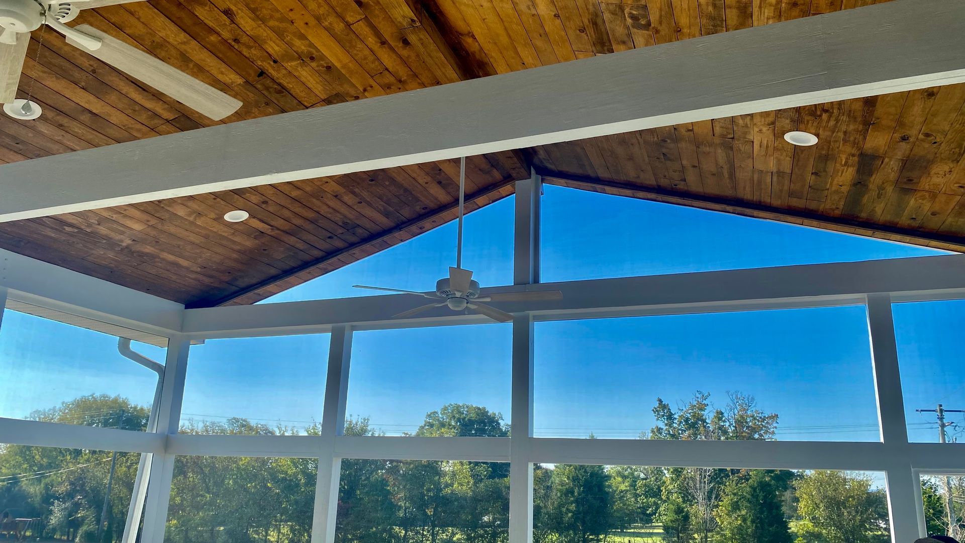 Screened-in porch with a wooden ceiling and large windows overlooking a sunny, tree-filled landscape.