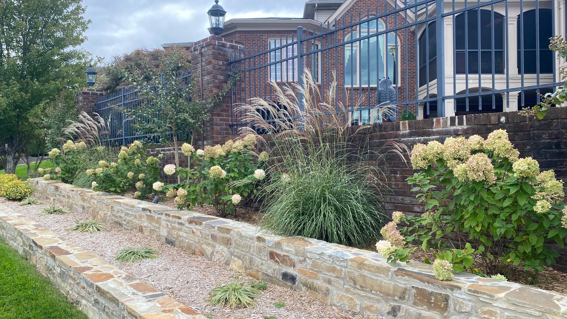 A brick retaining wall with flowering shrubs and ornamental grasses in front of a brick building with a wrought iron fence.