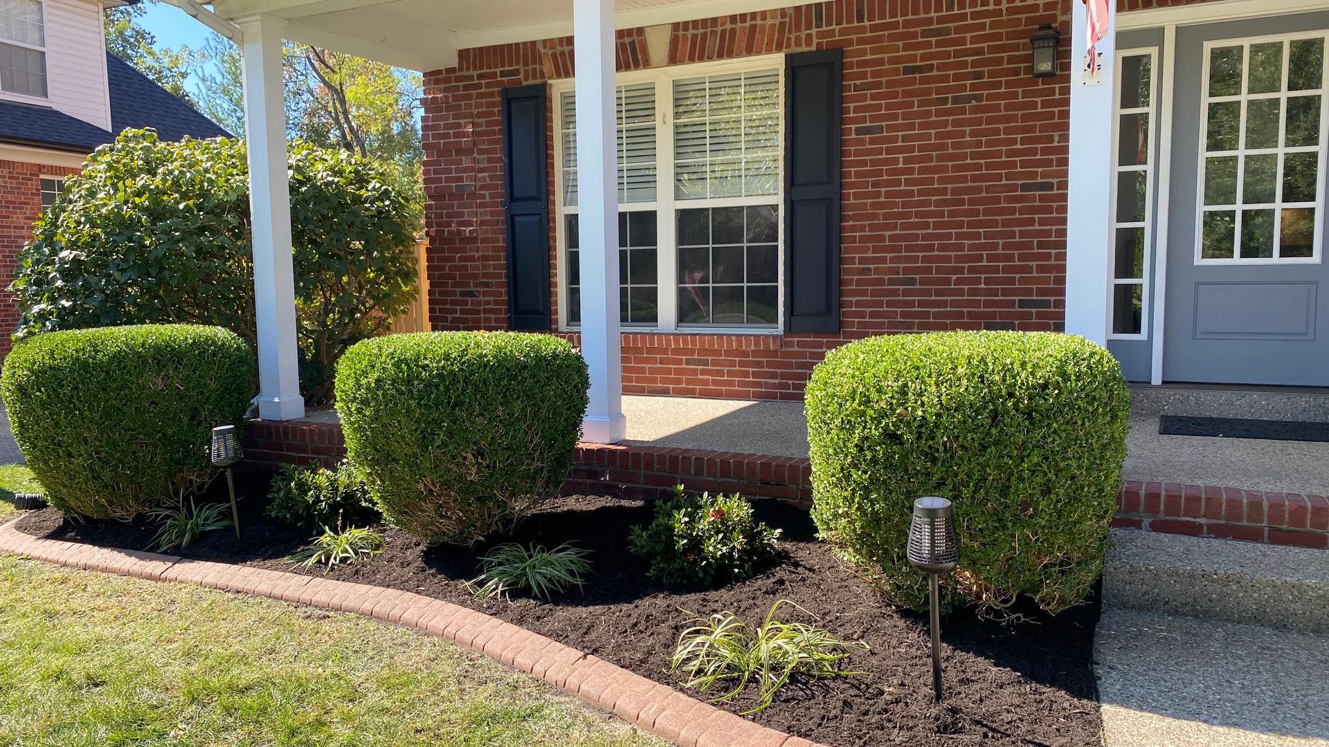 A flower bed with a brick border in front of a brick house. Mulch and young plants are visible.