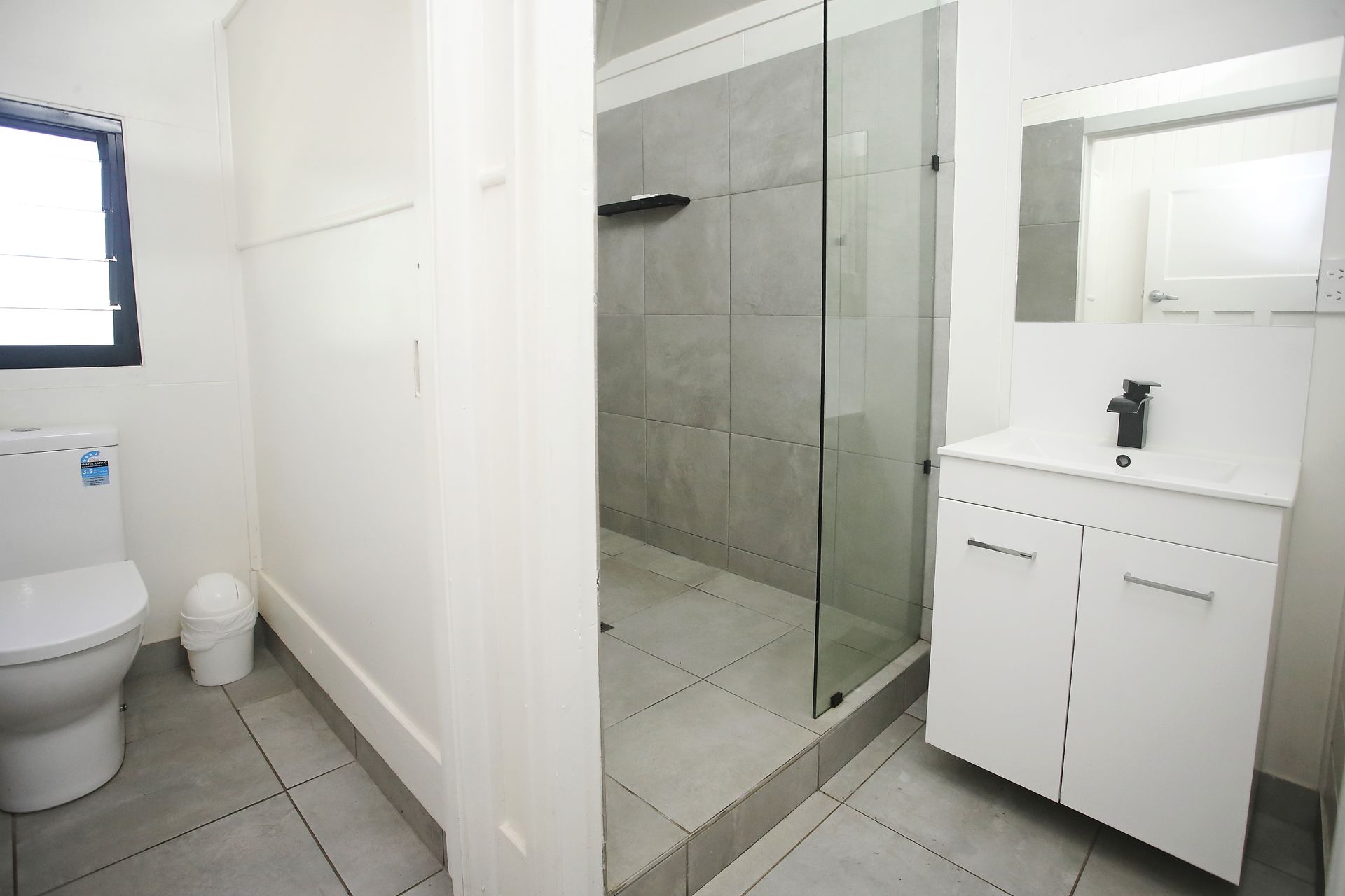 Bathroom with a toilet, vanity, and glass shower enclosure. White walls, grey tile flooring.