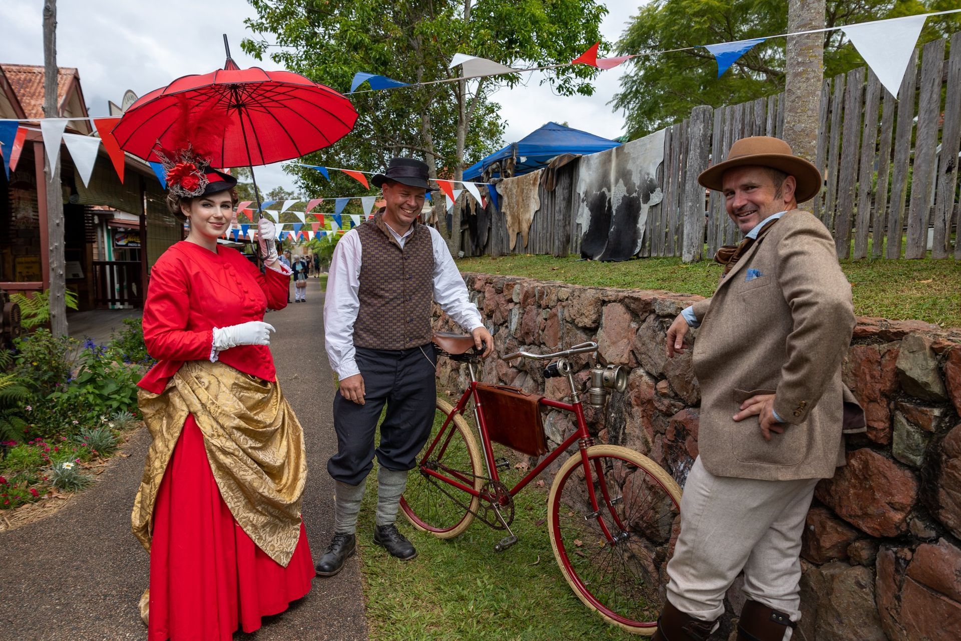 Three people in period clothing pose on a path. Woman in red dress with umbrella. Man with bike, other in tan coat.