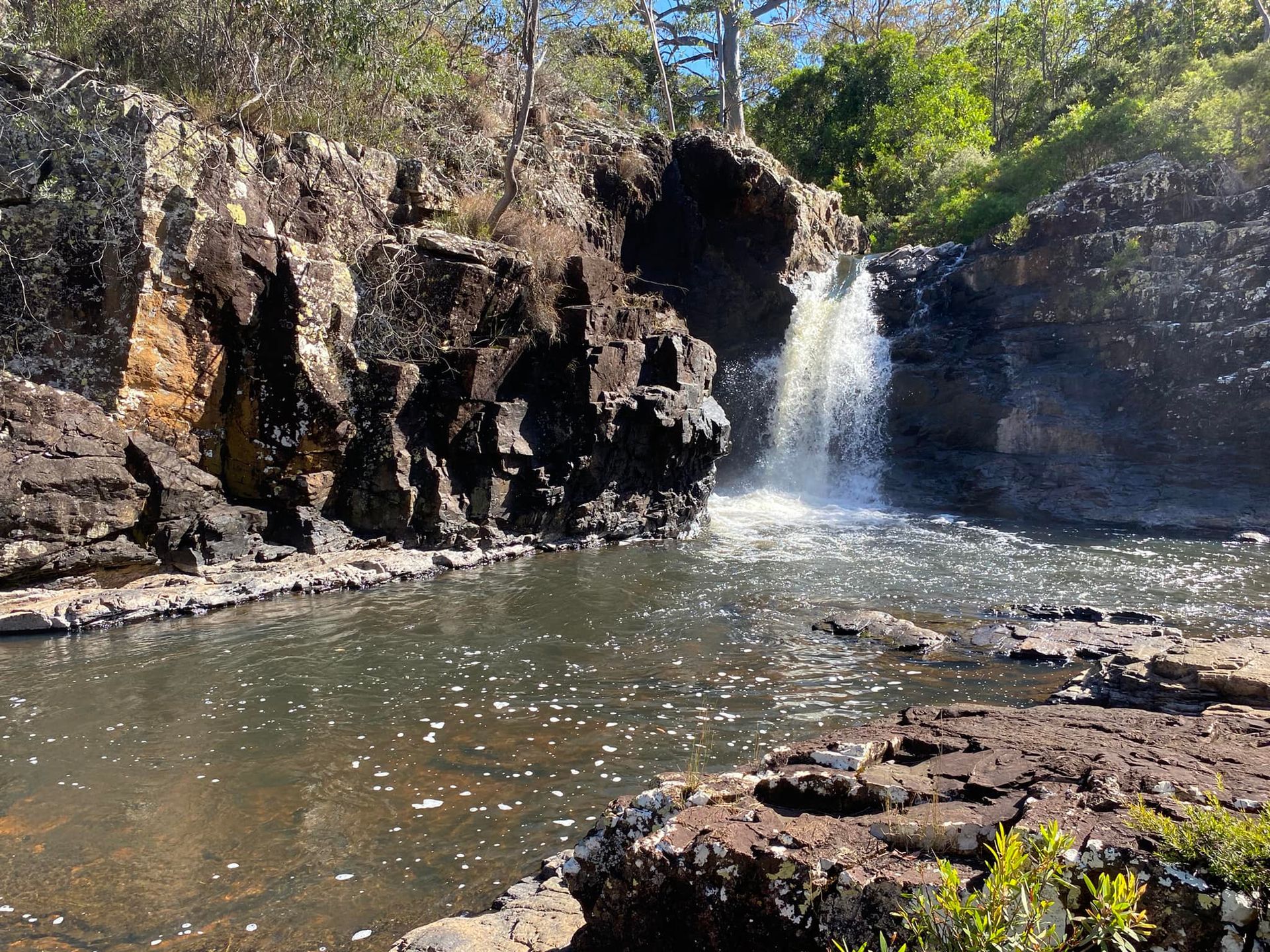 Waterfall cascading into a dark pool, surrounded by dark rocks and sparse green vegetation.
