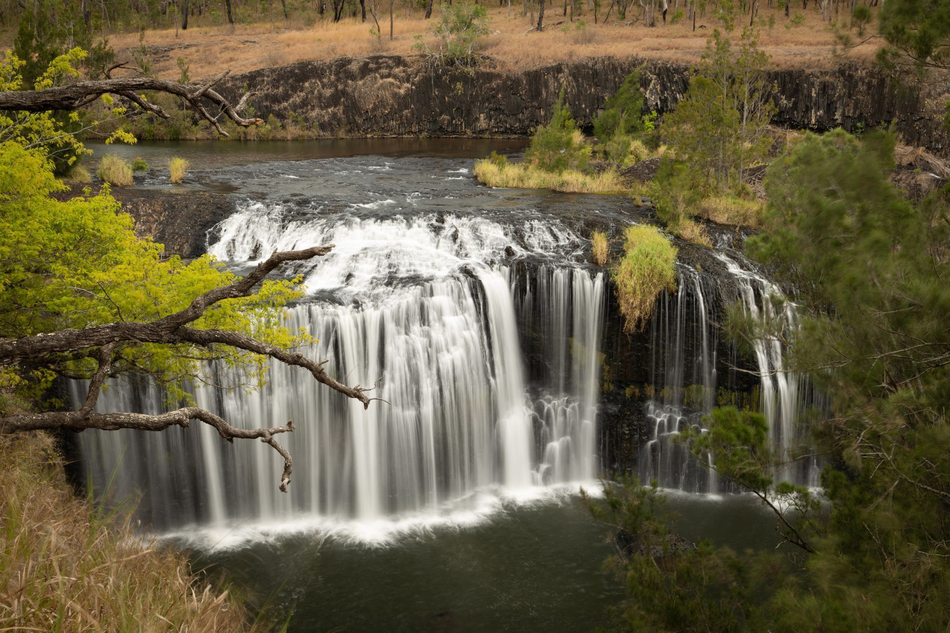 Waterfall cascading over dark rocks into a pool, surrounded by green foliage and trees.