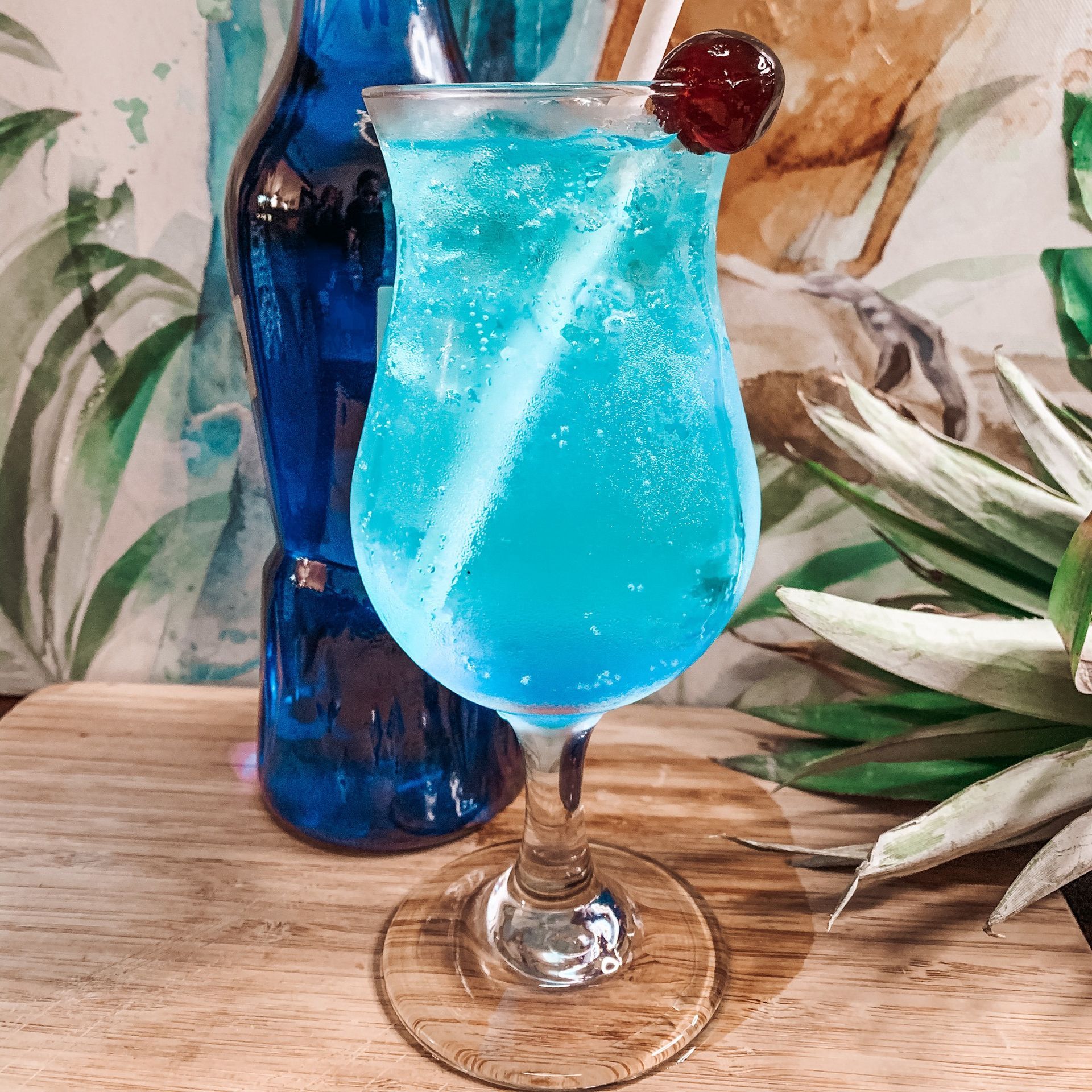 Blue cocktail in a stemmed glass, cherry garnish, blue bottle background.