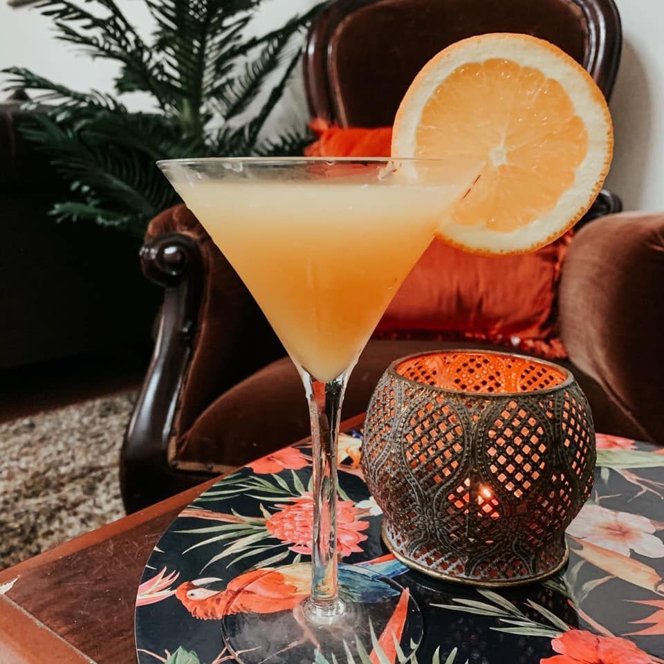 Cocktail in a martini glass with orange slice garnish, on a table with a lit candle and floral placemat.