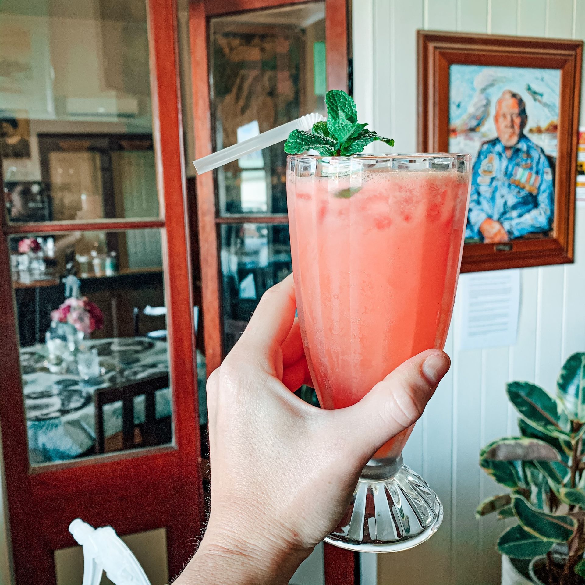 Hand holding a pink cocktail with mint garnish in a tall glass, near a portrait and a plant, in a cafe setting.