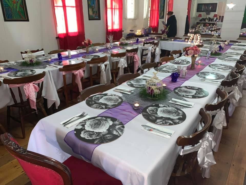 Tables set for a gathering, white tablecloths with purple runners. Pink accents on chairs. Indoors, natural light.