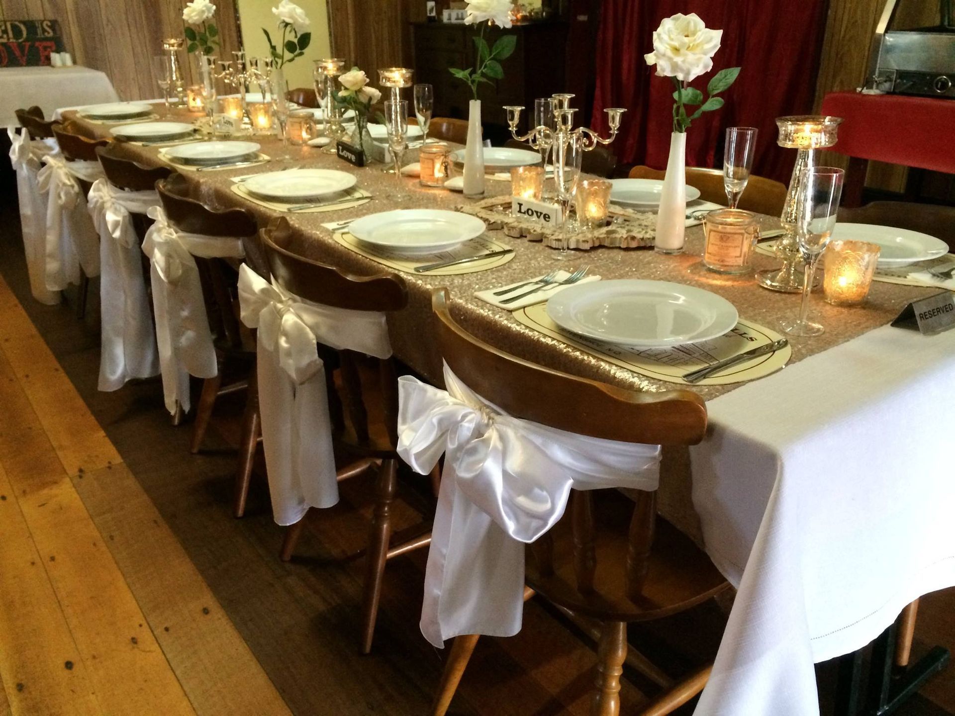 Long dining table set for a wedding with white tablecloth, gold sequined runner, floral centerpieces, and white bow chair decor.
