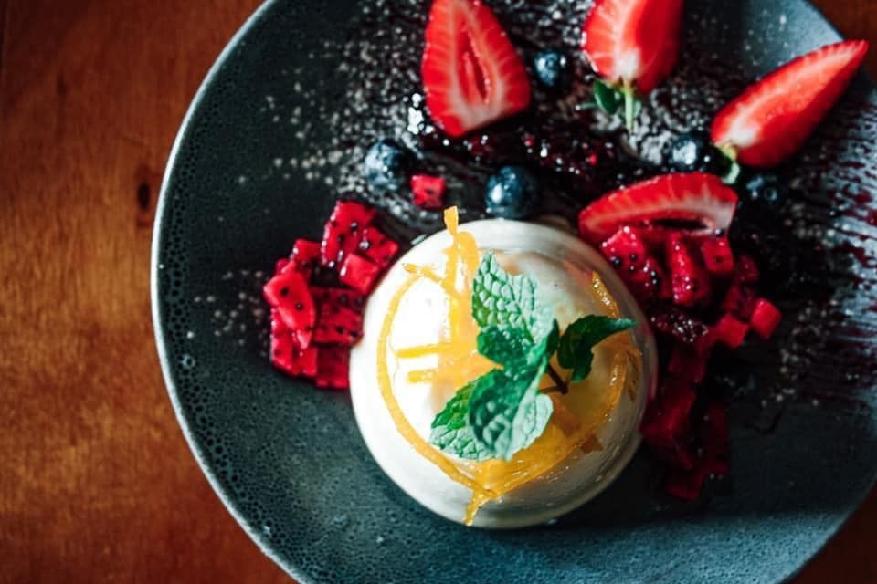 Dessert with white cream, fresh strawberries, blueberries, and chopped red fruit on a dark plate.