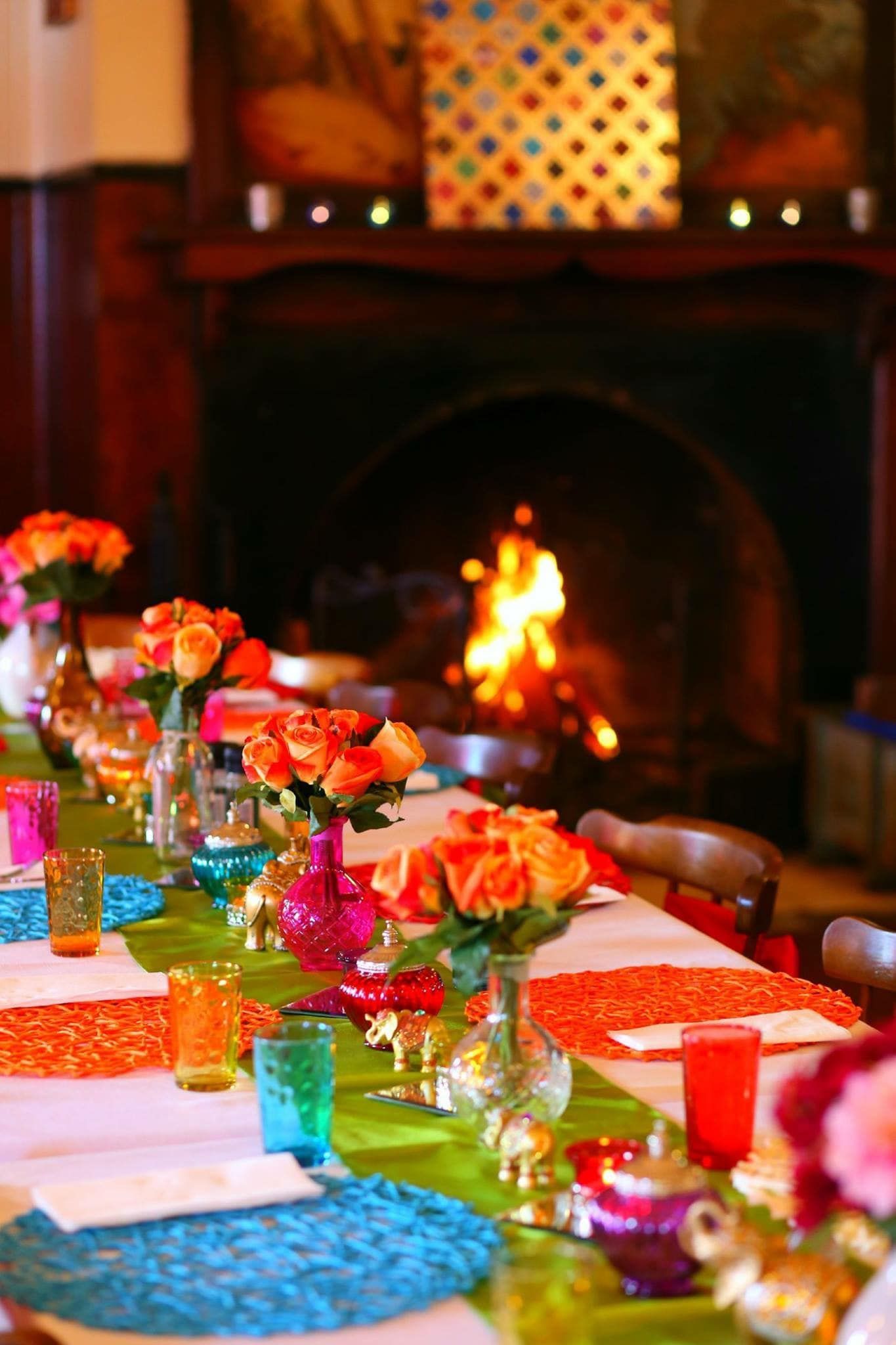 Long table set for a party with bright orange flowers, colorful place settings, and a fireplace in the background.