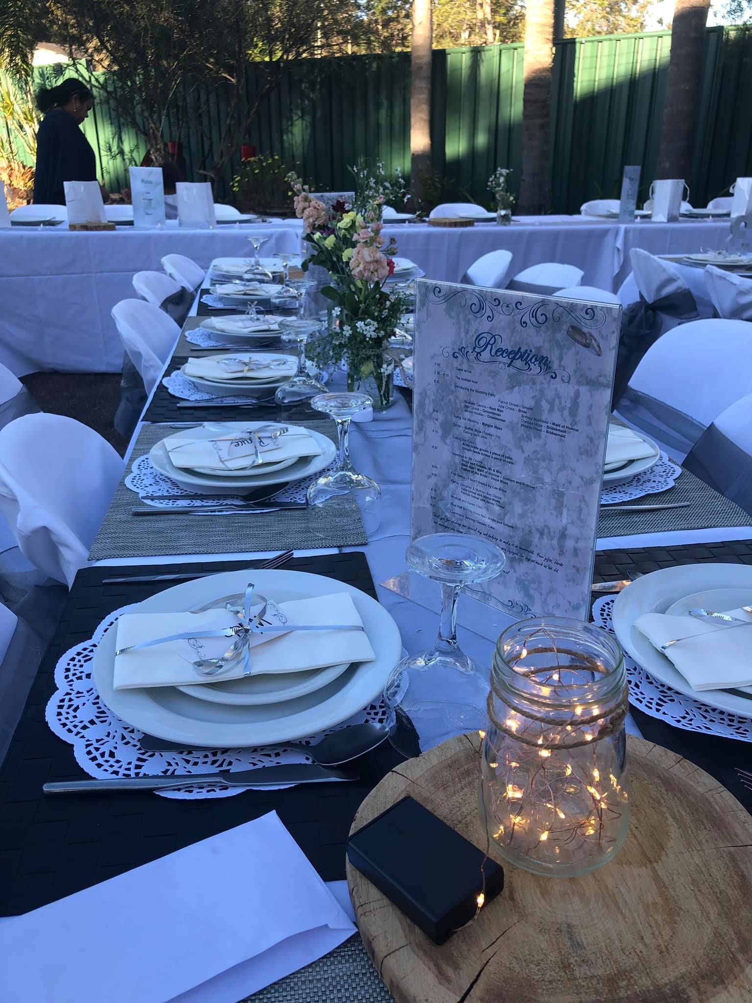 Outdoor tables set for a gathering with white plates, flowers, and a centerpiece with fairy lights.