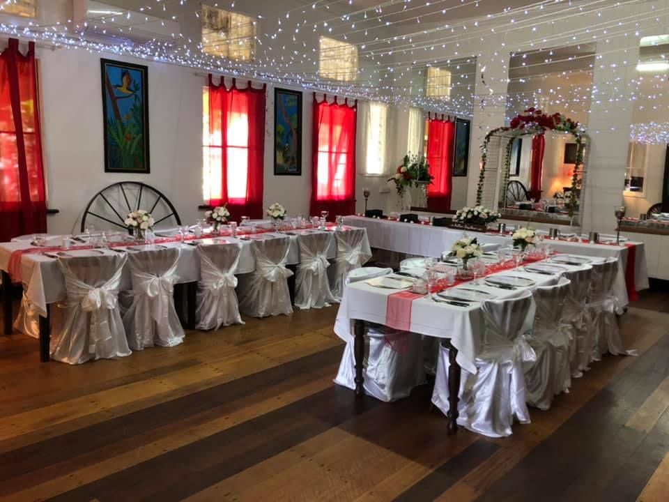 Formal dining room set for an event; tables with white linens, red accents, decorated chairs, wood floor.