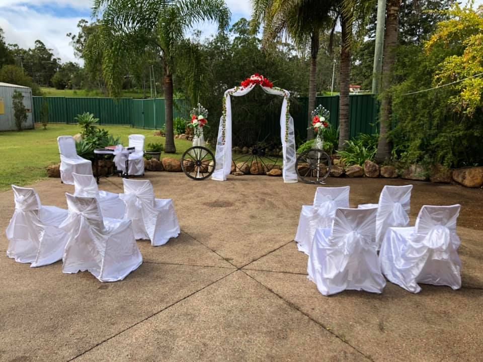 Outdoor wedding setup with white-covered chairs facing an archway with floral decorations; green yard backdrop.