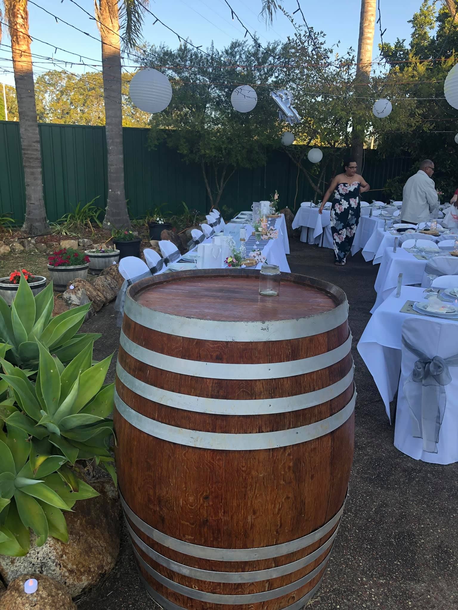 Wooden barrel in the foreground, with tables set for an outdoor event in a garden, with guests and decorations.