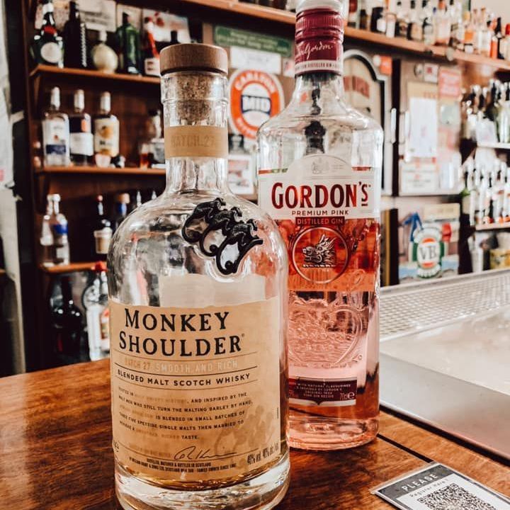 Monkey Shoulder whisky bottle and Gordon's pink gin bottle on a bar top, liquor shelves in background.