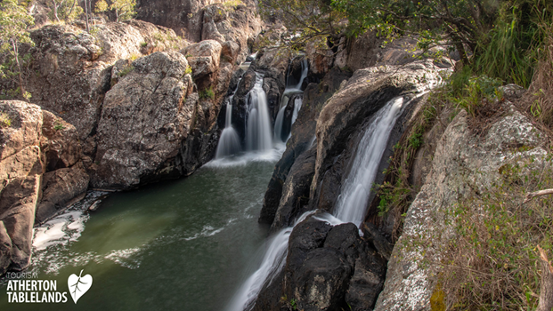 Water cascades over multiple waterfalls into a green pool, surrounded by rocks and vegetation.